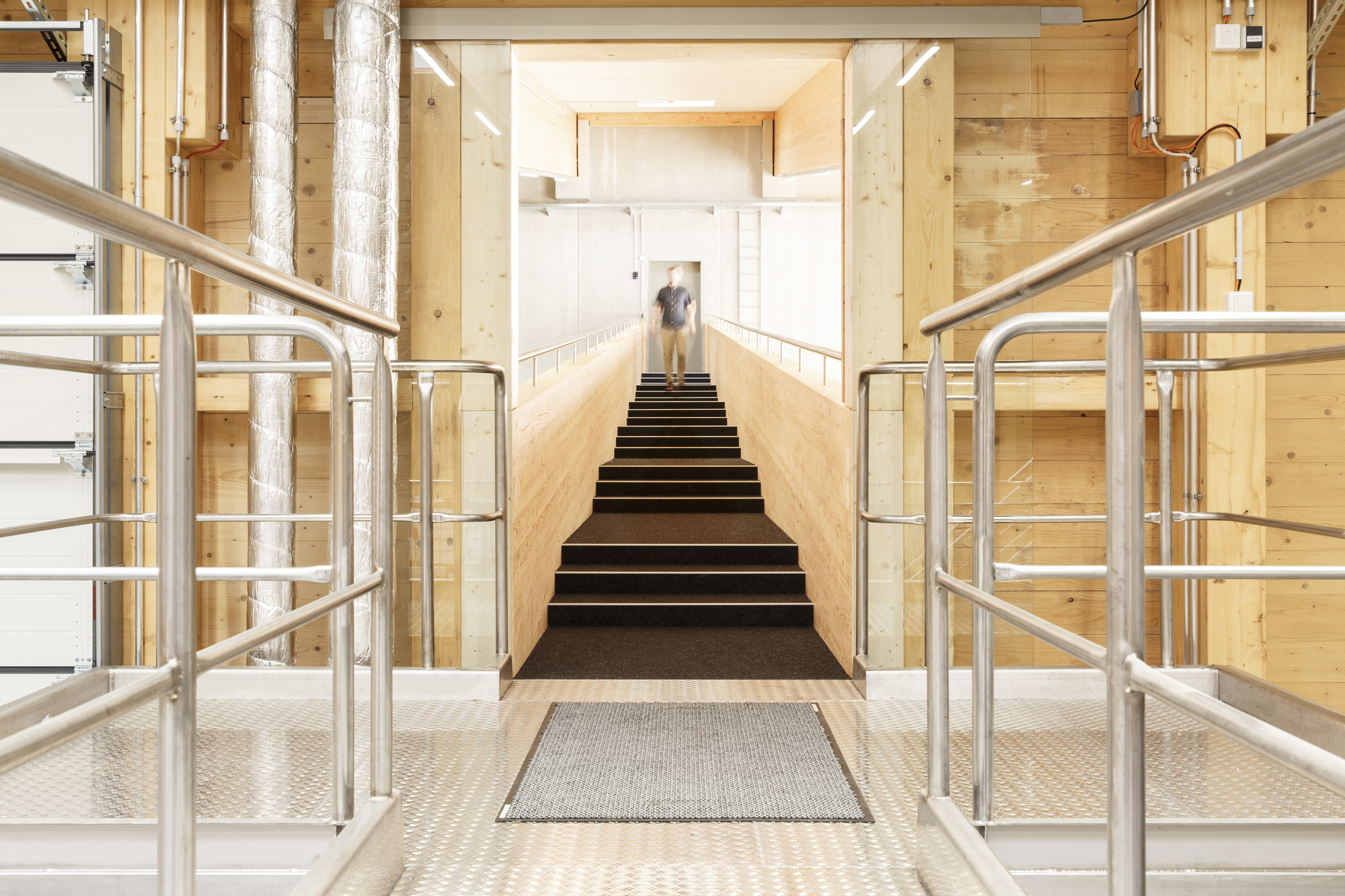 Central stairway flanked by steel railings and exposed timber-lined walls with a person at the landing