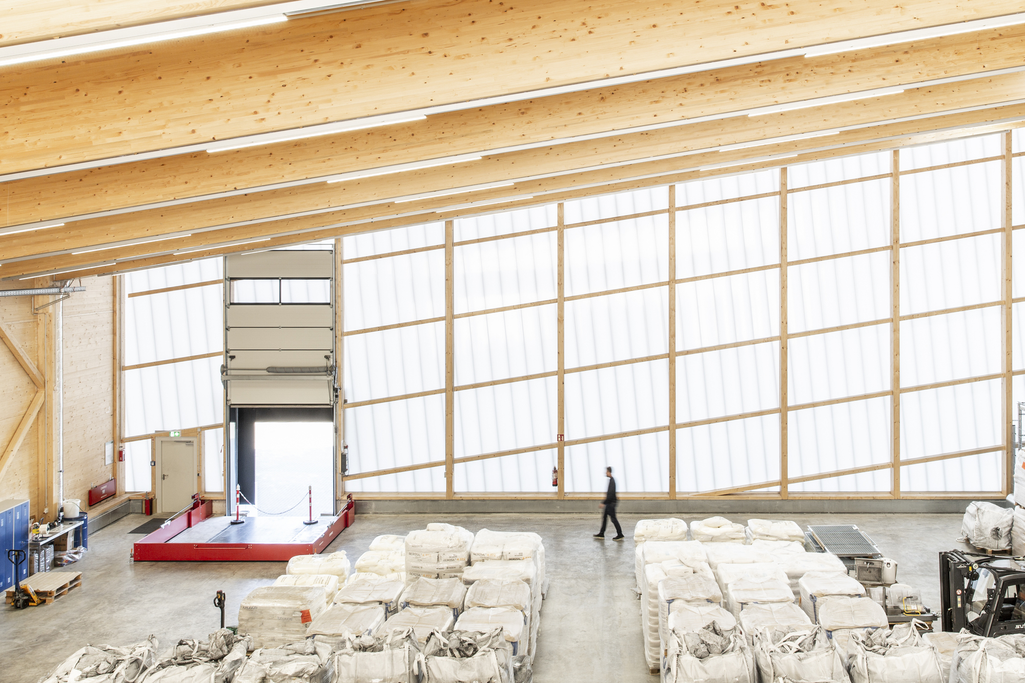 High-ceilinged production hall with translucent corrugated wall panels, exposed timber beams, and a person walking
