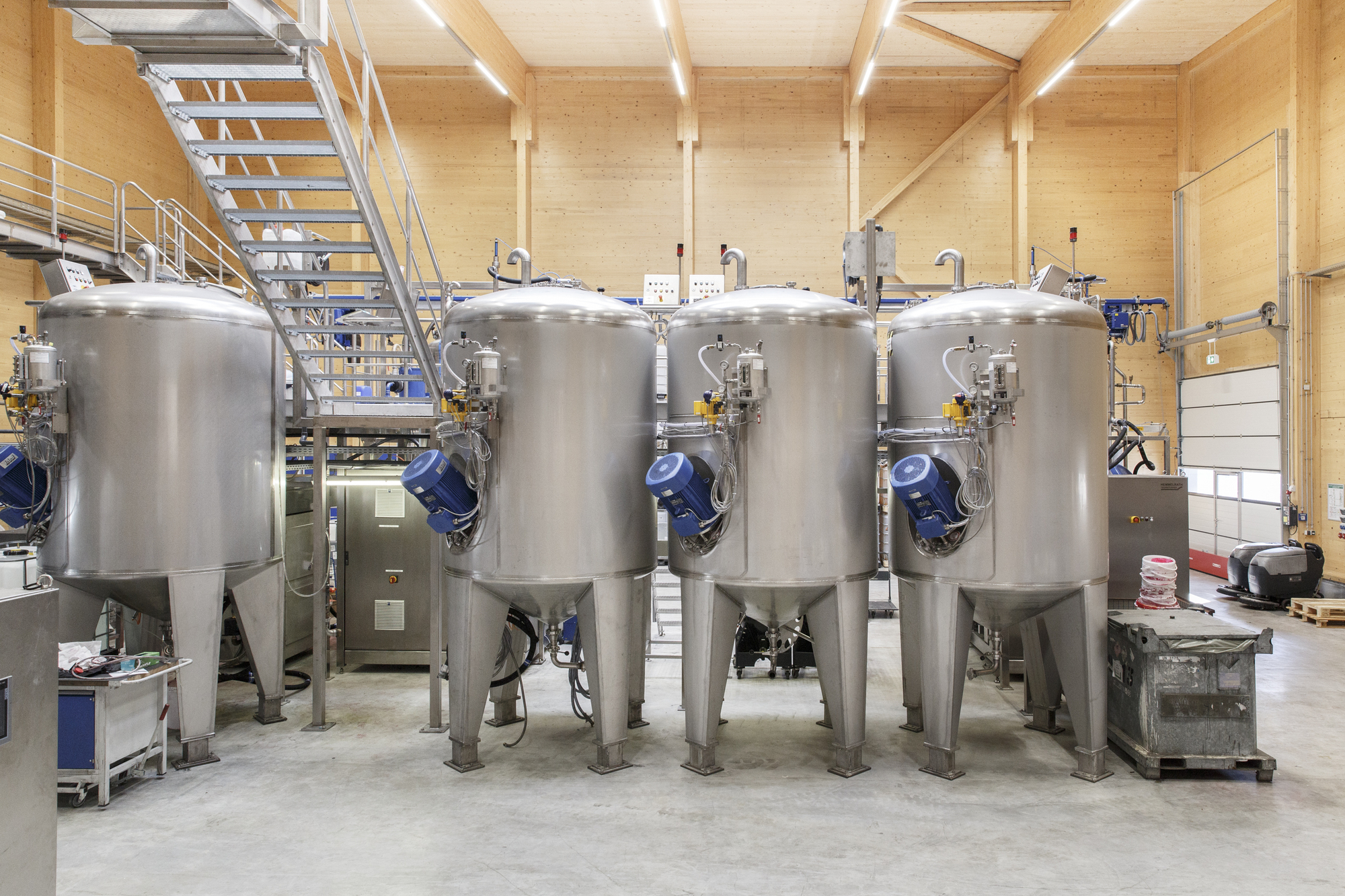 Row of stainless steel fermentation tanks with blue valves on a concrete floor beneath exposed timber beams