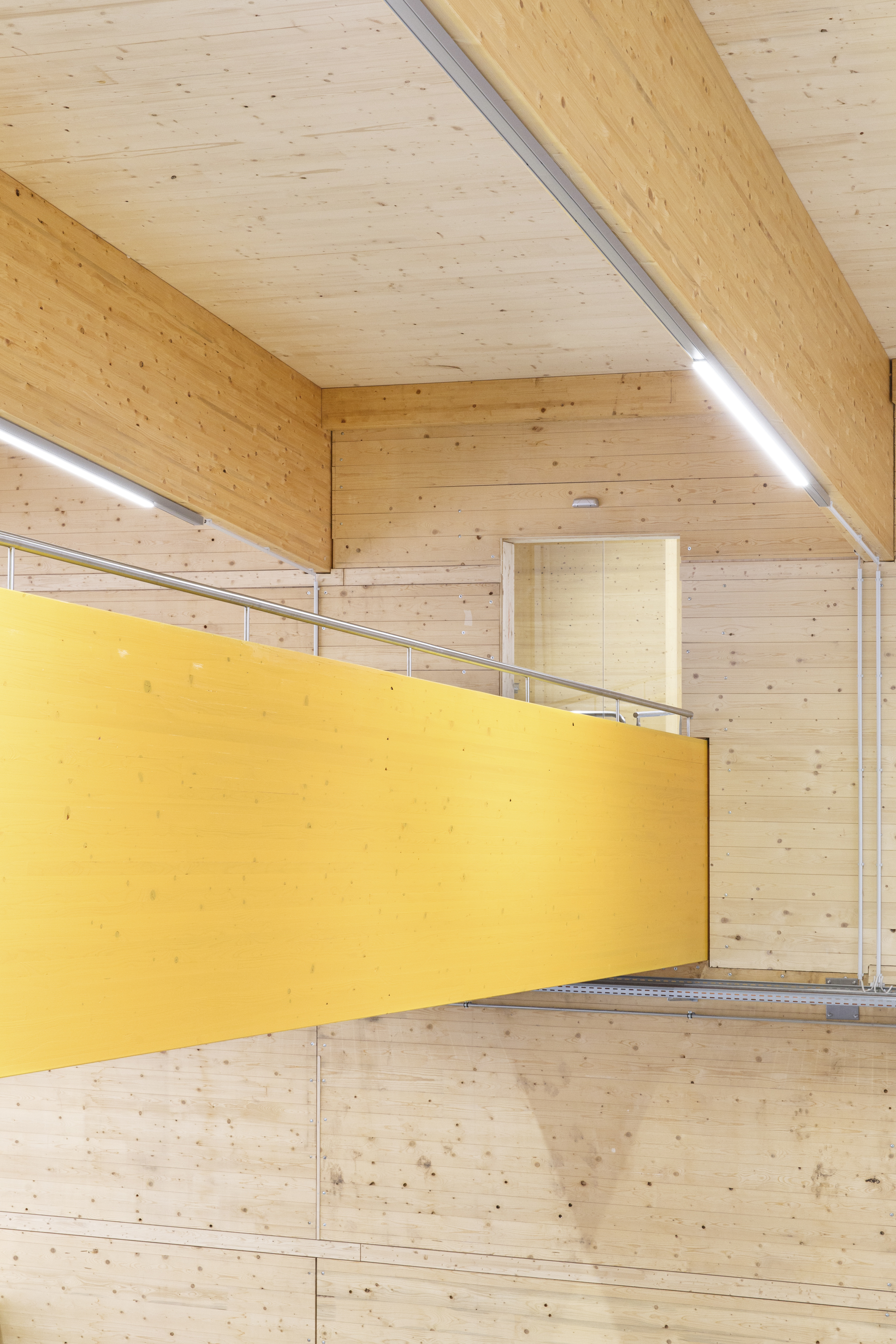 Yellow mezzanine walkway with metal railing suspended within double-height space clad in exposed timber planks