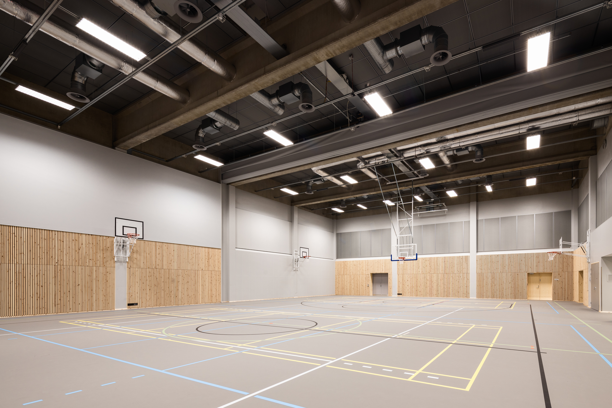 Sports hall with marked floor, retractable basketball hoops and timber wainscoting below exposed mechanical ceiling