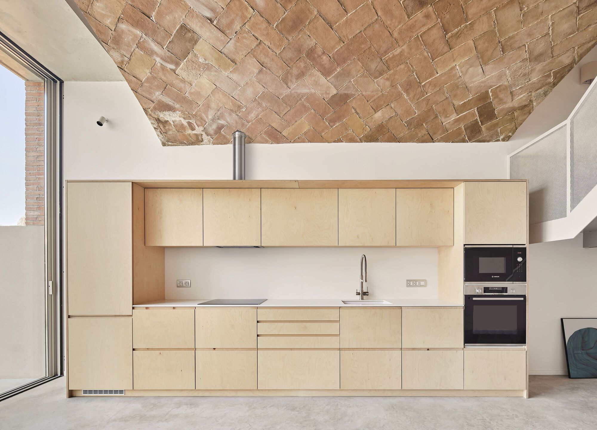 Light plywood kitchen cabinetry under a herringbone brick vault with a steel flue pipe rising through