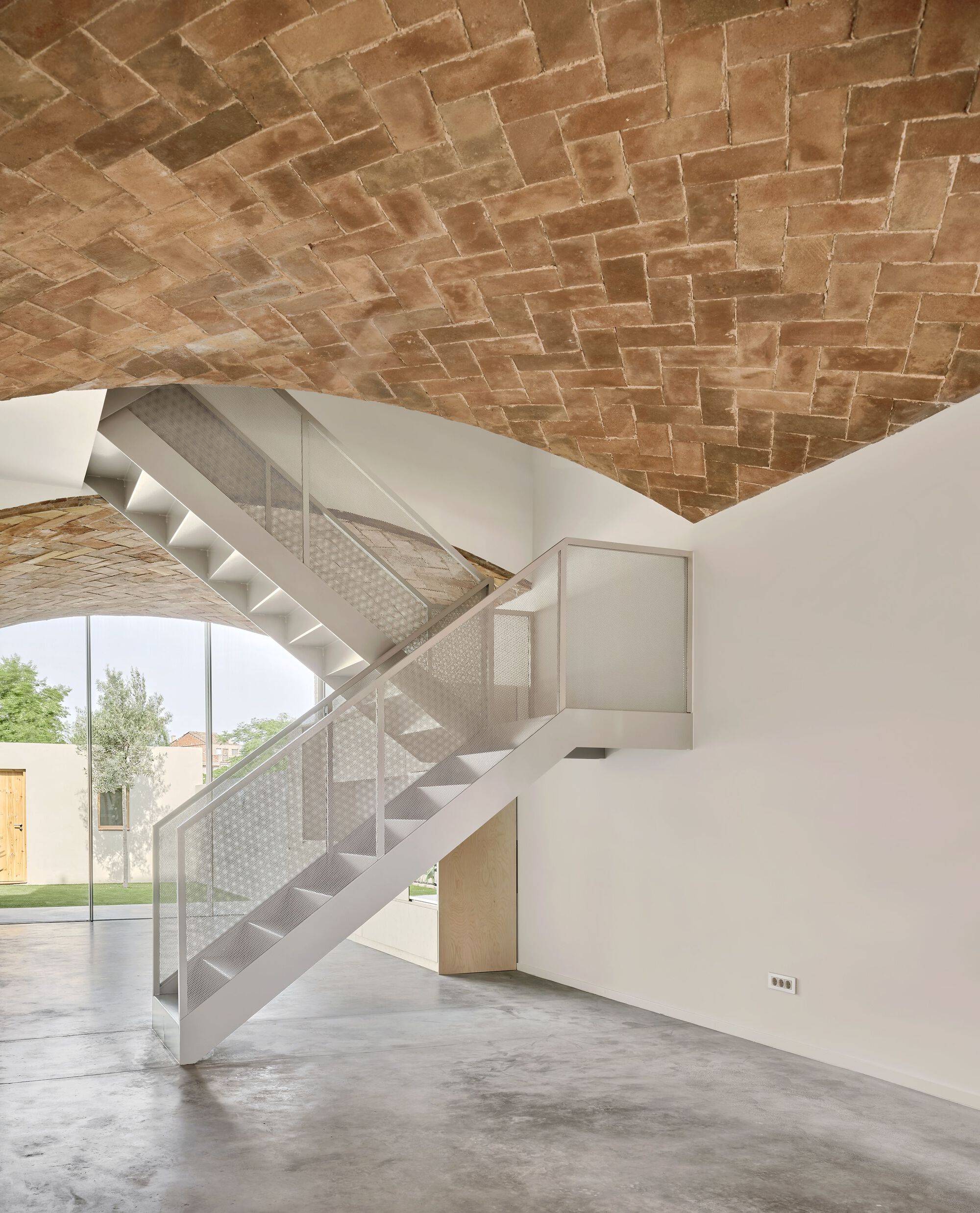 Perforated metal staircase beneath a herringbone brick vault with garden visible through glazed opening
