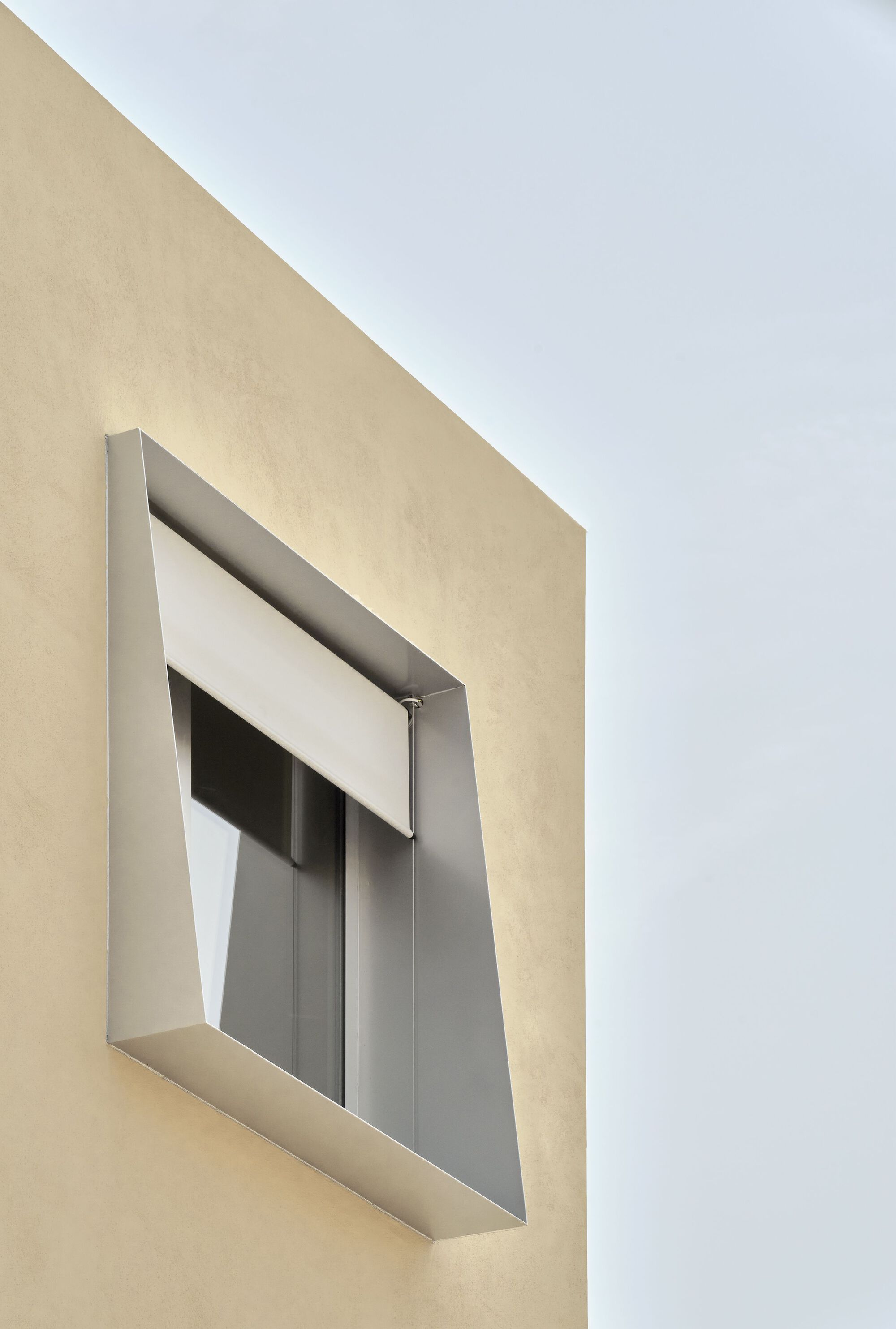 Close-up of recessed window opening with angled metal frame and roller shutter on cream facade