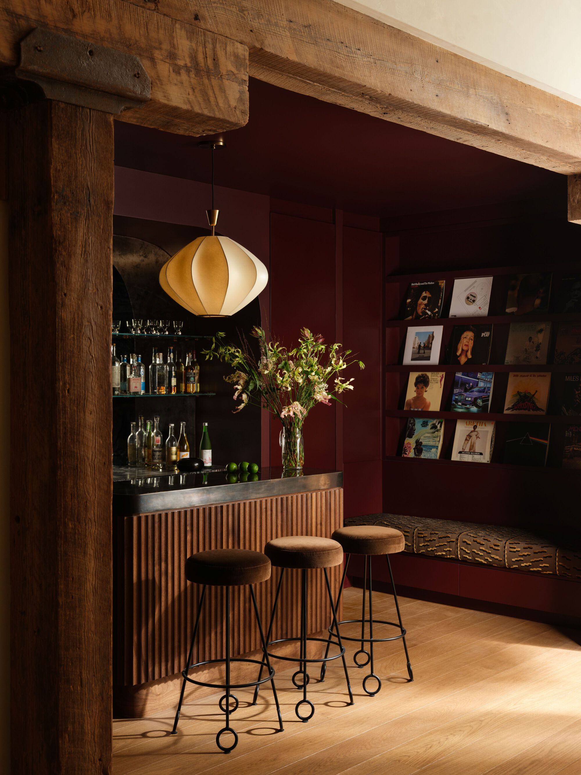 Bar nook with fluted timber front, burgundy walls, record display, and pendant light under timber beam