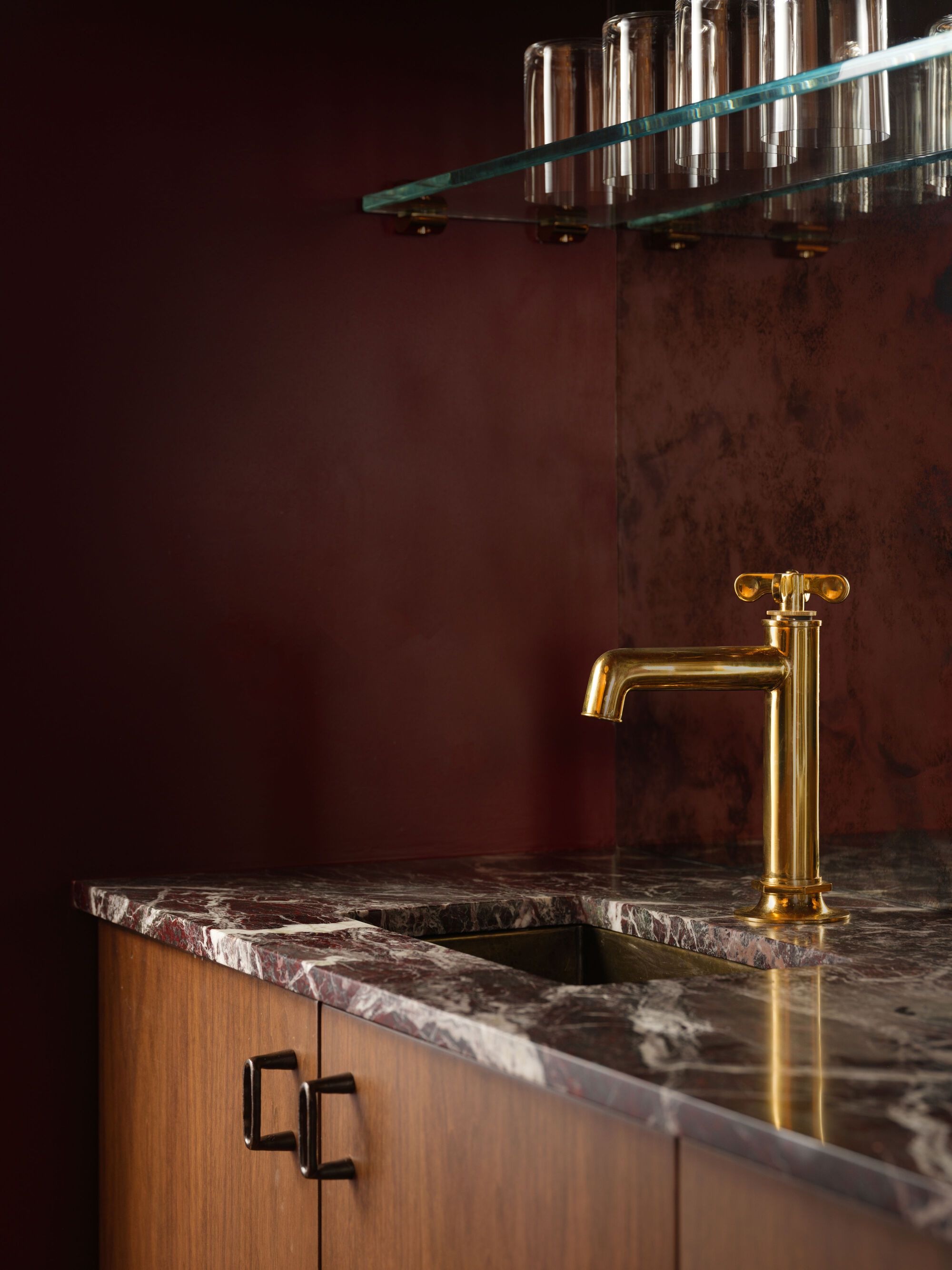 Brass faucet on burgundy veined marble counter against oxidized copper wall panel with glass shelf