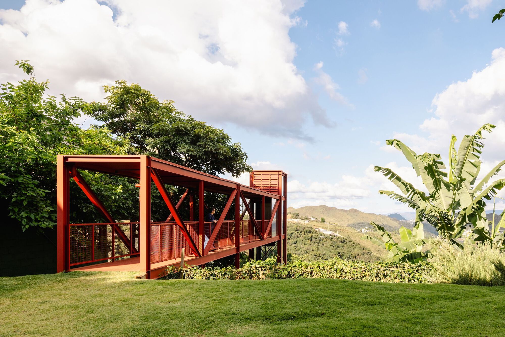 Red steel-framed pavilion with diagonal bracing set on a sloped lawn with banana plants and mountain backdrop