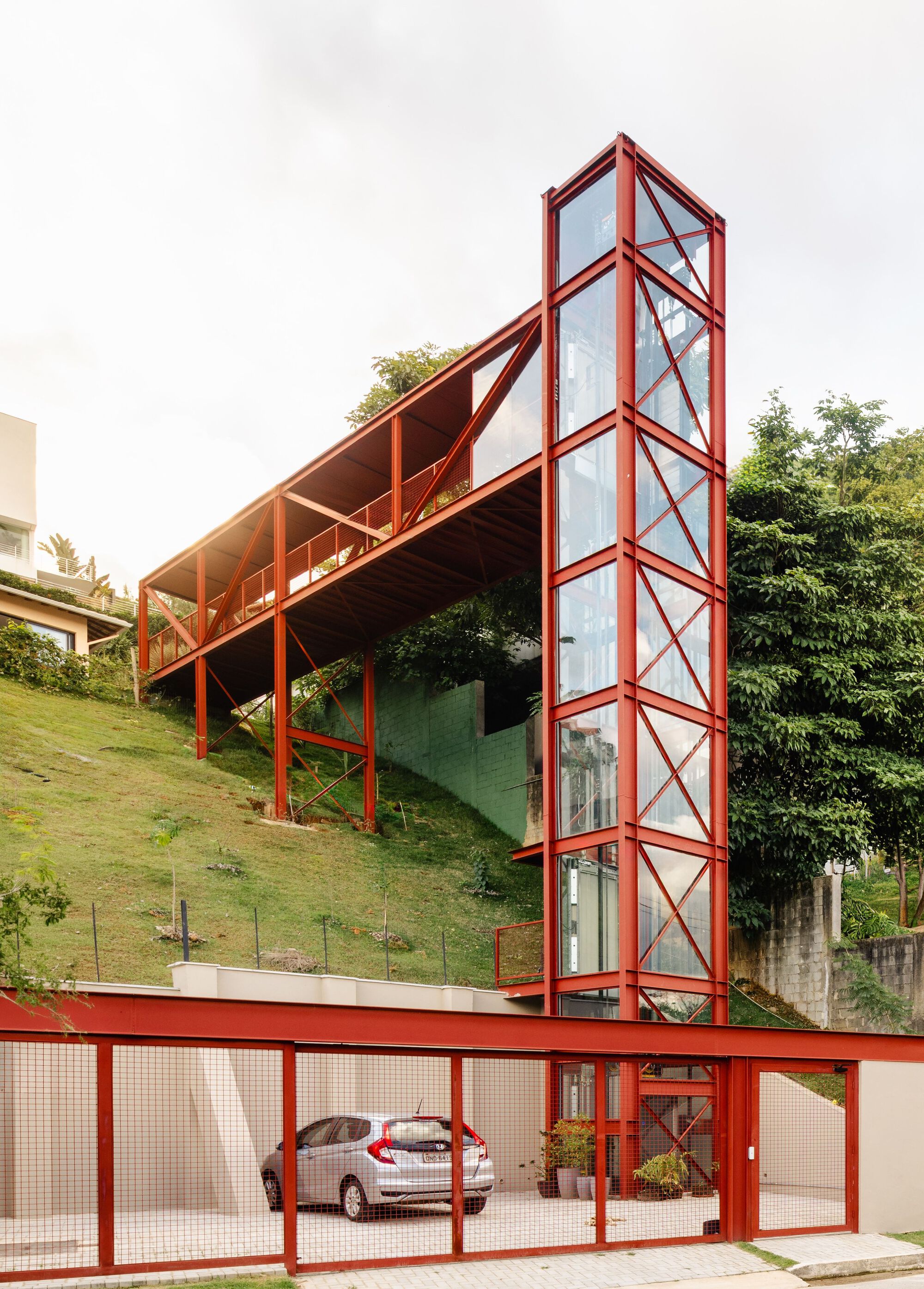 Vertical glass circulation tower clad in red steel with diagonal cross-bracing rising from a covered carport