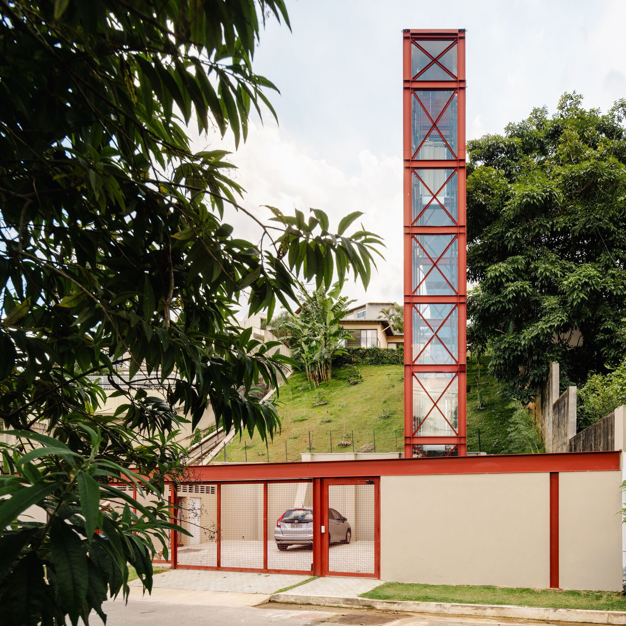 Street facade with red steel frame and mesh gate enclosing a covered carport under a sloped lawn