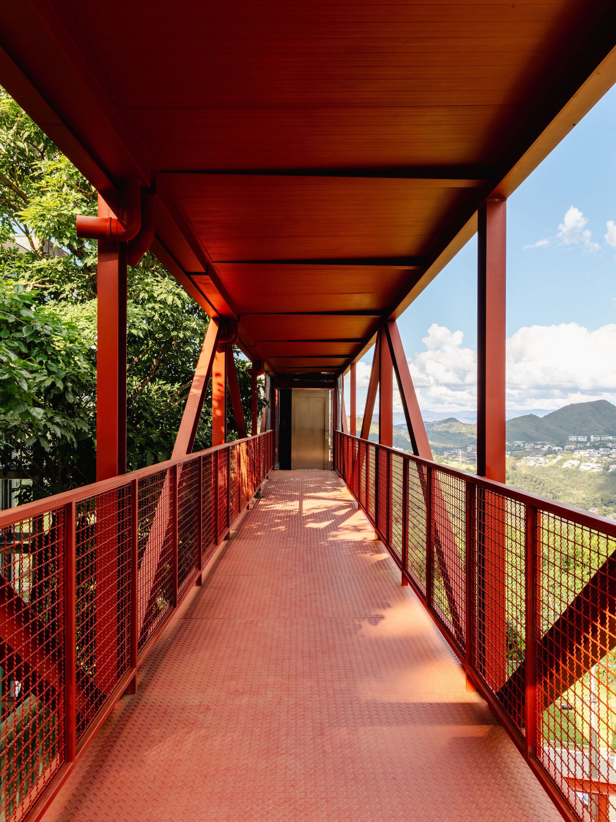 Covered walkway with red steel structure, mesh safety railings, and metal grate flooring looking toward mountain views