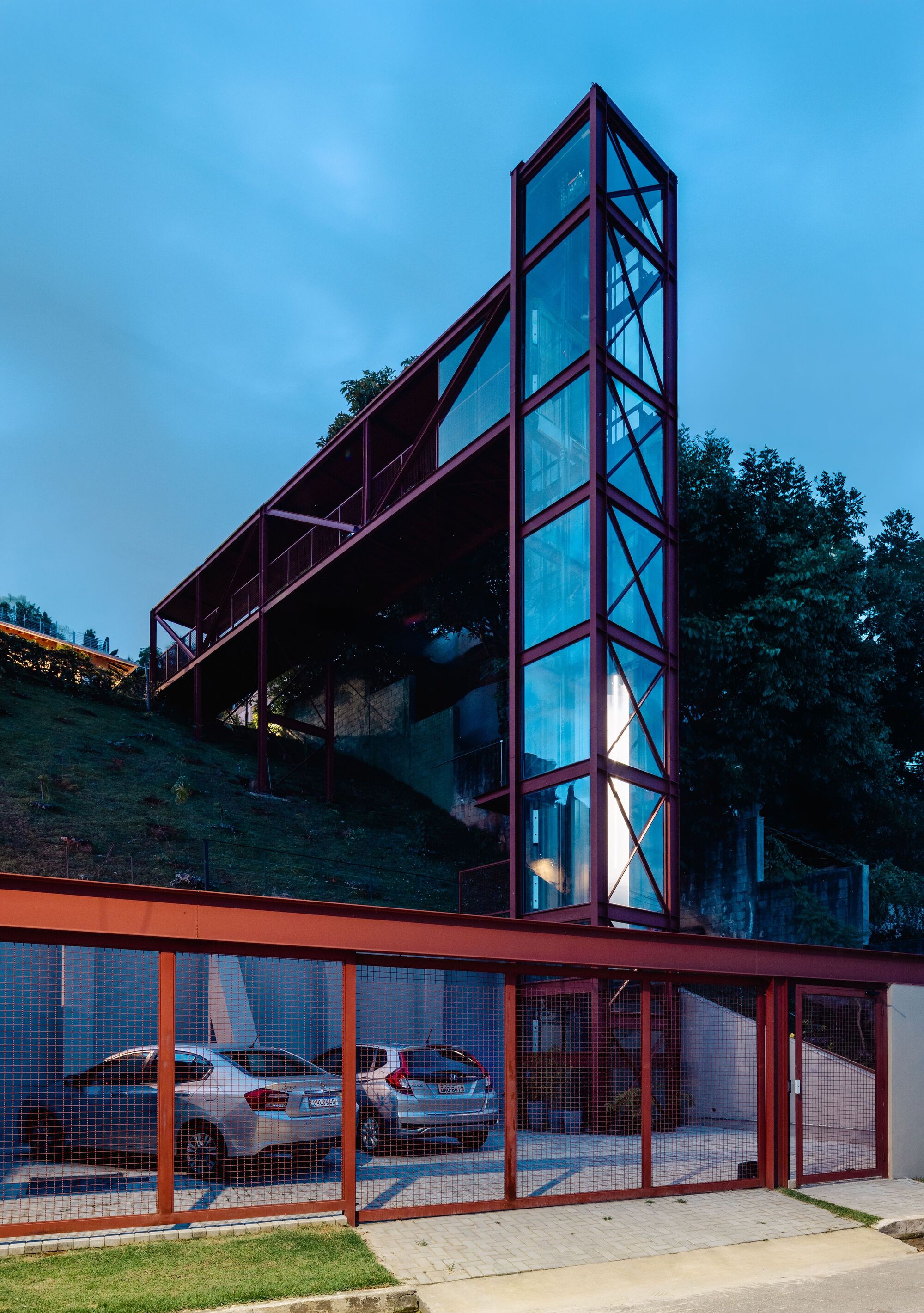 Red steel structure with glazed elevator tower rising above a mesh-enclosed carport at dusk