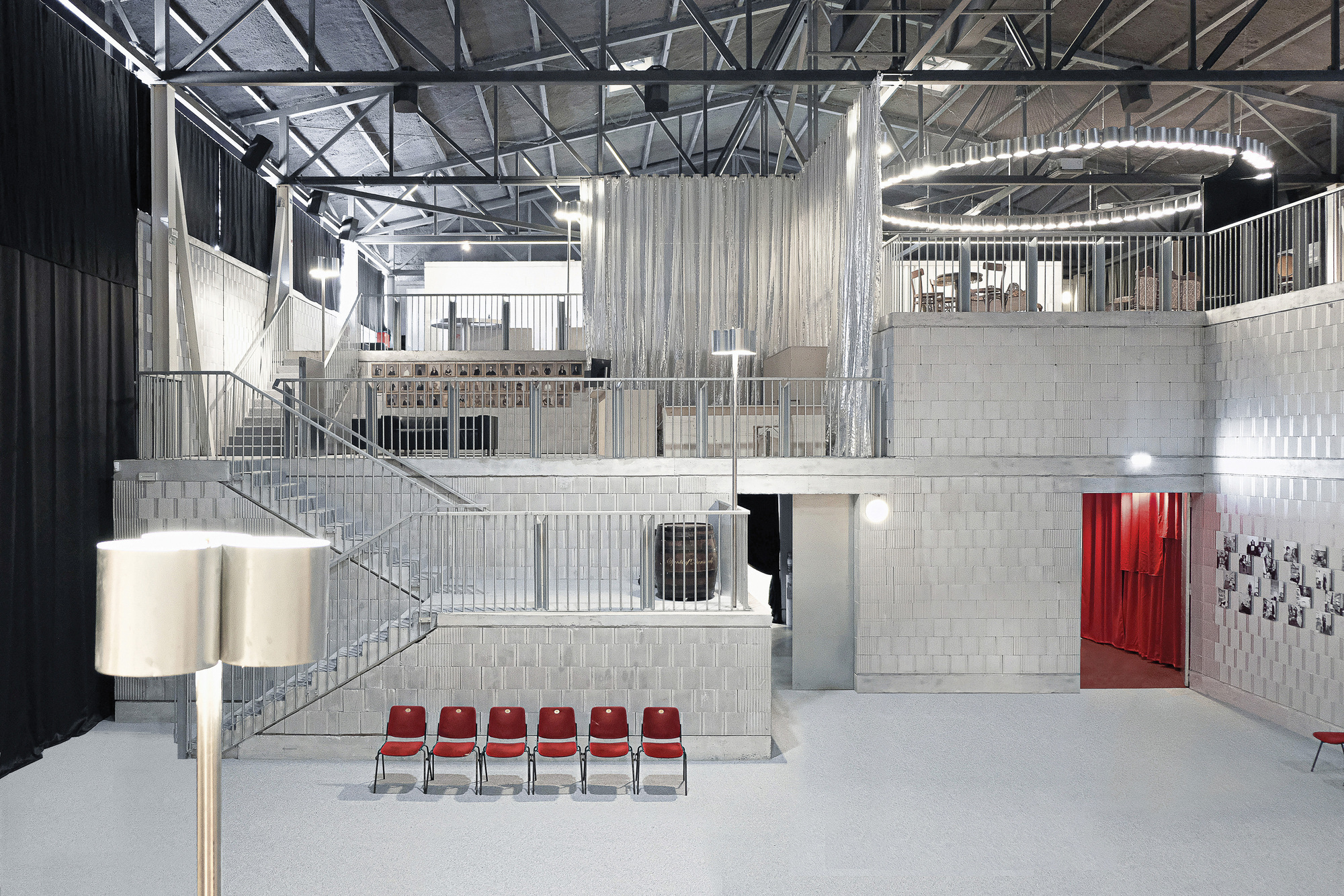 Triple-height interior hall with exposed steel trusses, metal railings, concrete platforms and row of red chairs