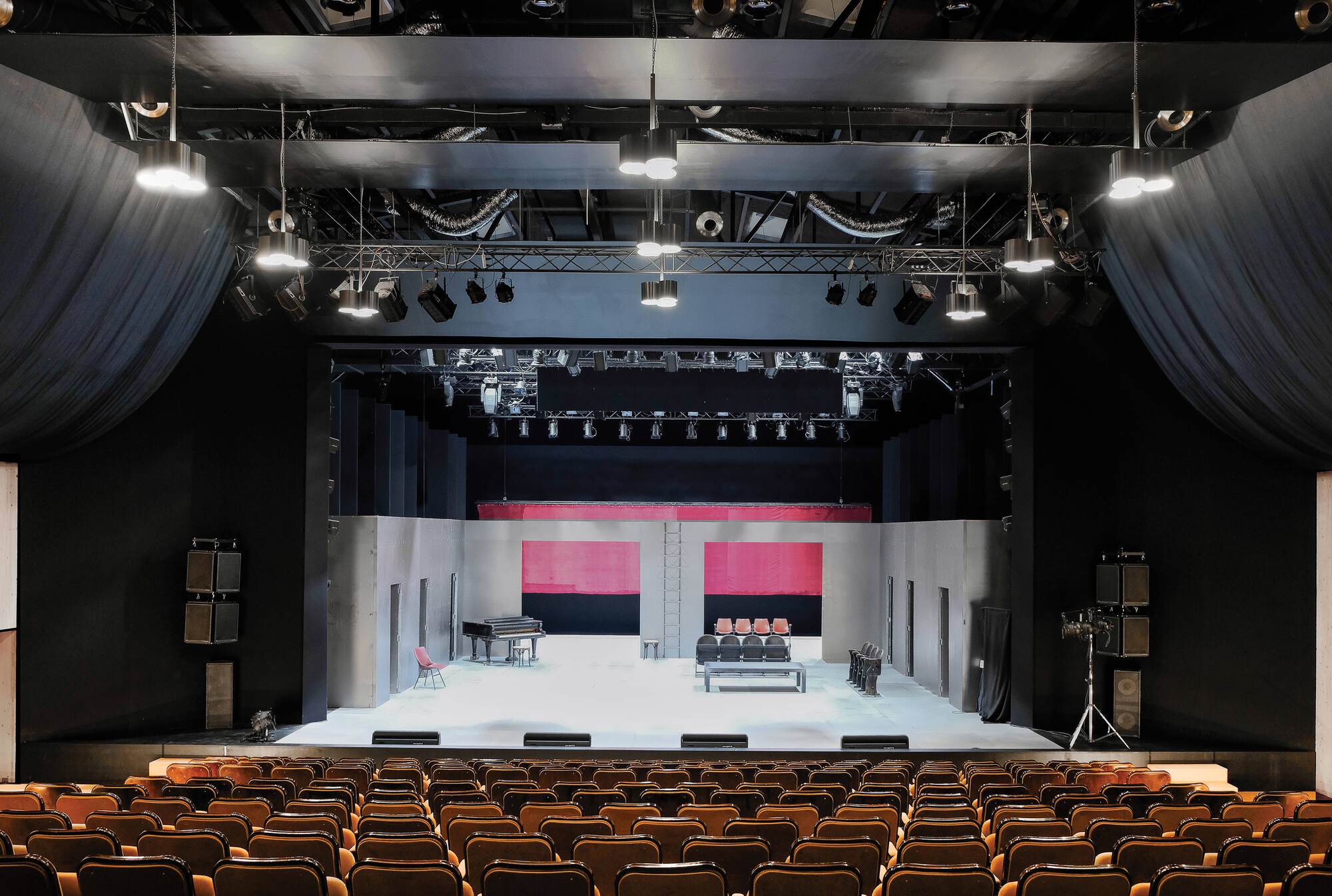 Theater auditorium with exposed black grid, stage lighting, rows of upholstered seats, and a white set