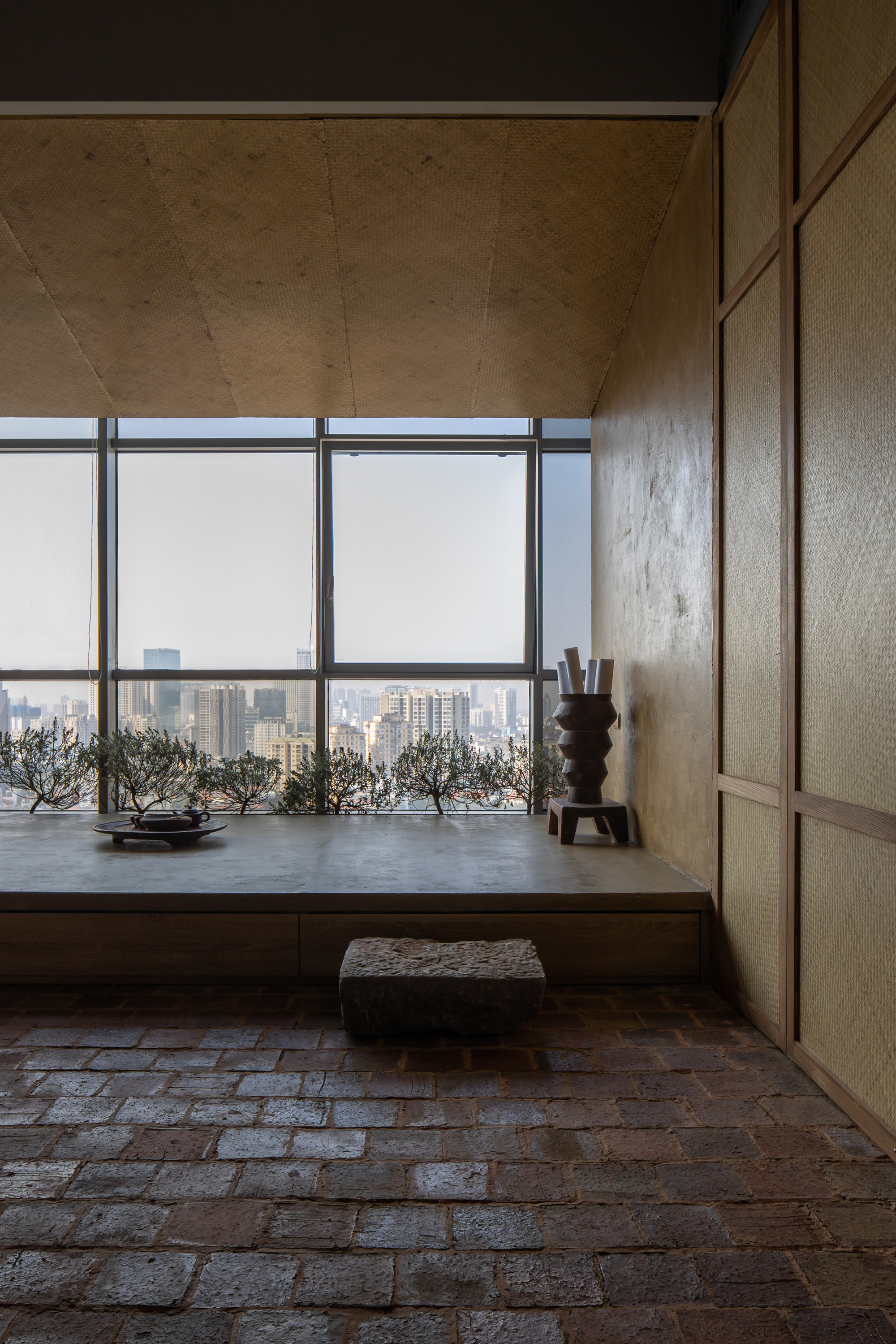 Raised timber platform with potted bonsai trees and floor cushion overlooking hazy cityscape through full-height glazing