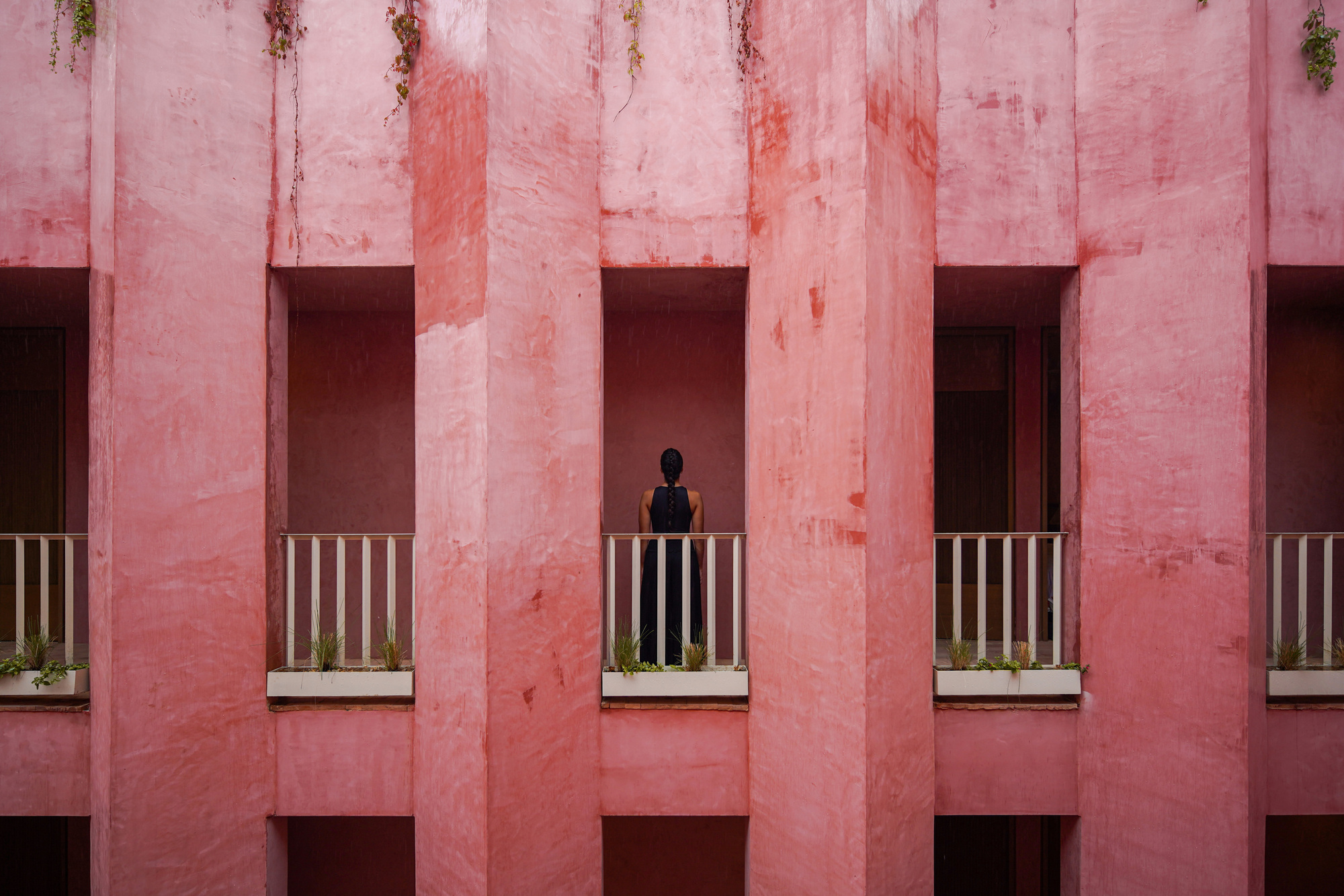 Facade detail with vertical pink concrete fins and a figure standing on a balcony with striped railings