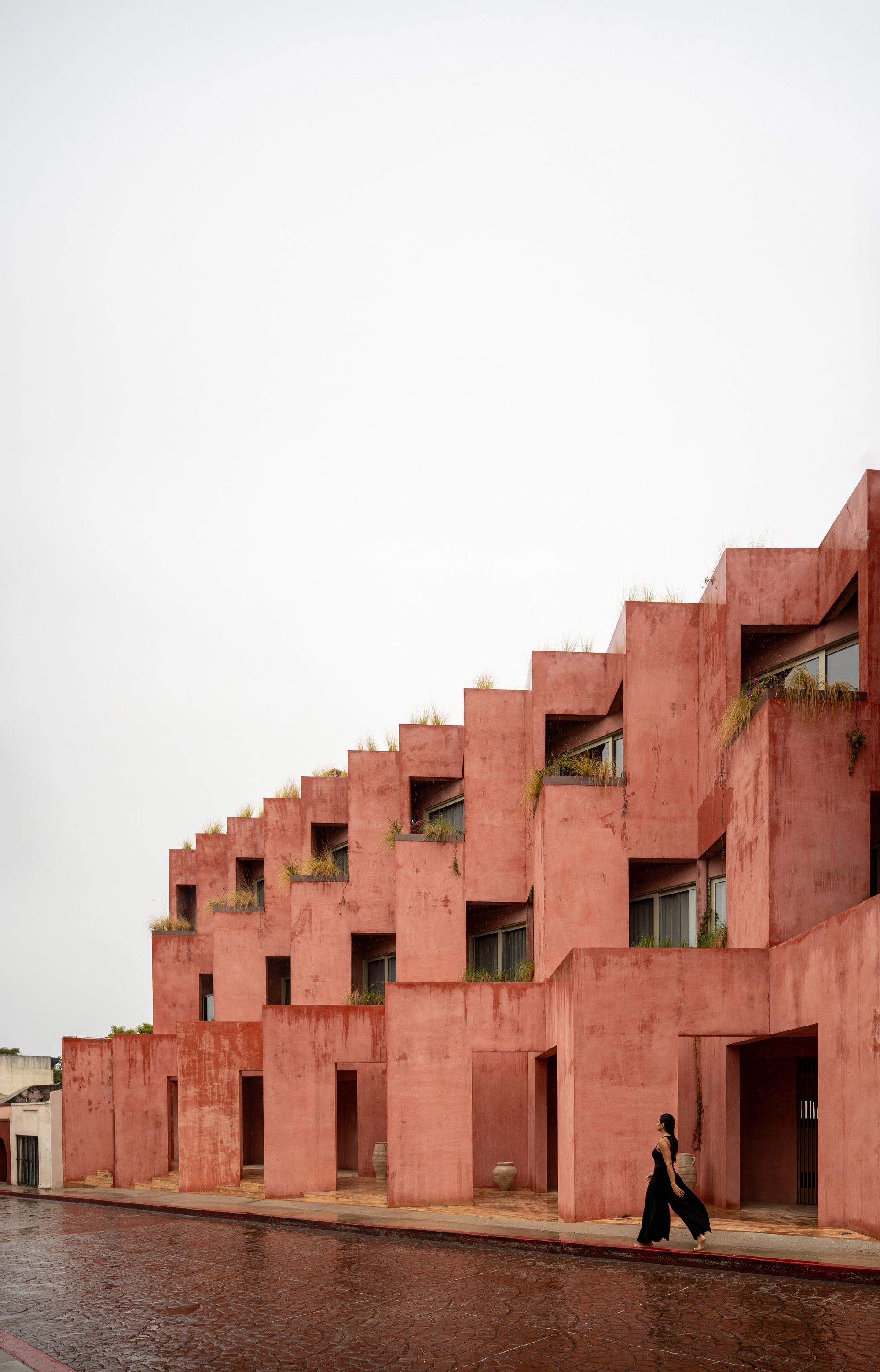 Stepped red plaster facade with planted terraces and deep-set openings as a person walks on wet pavement