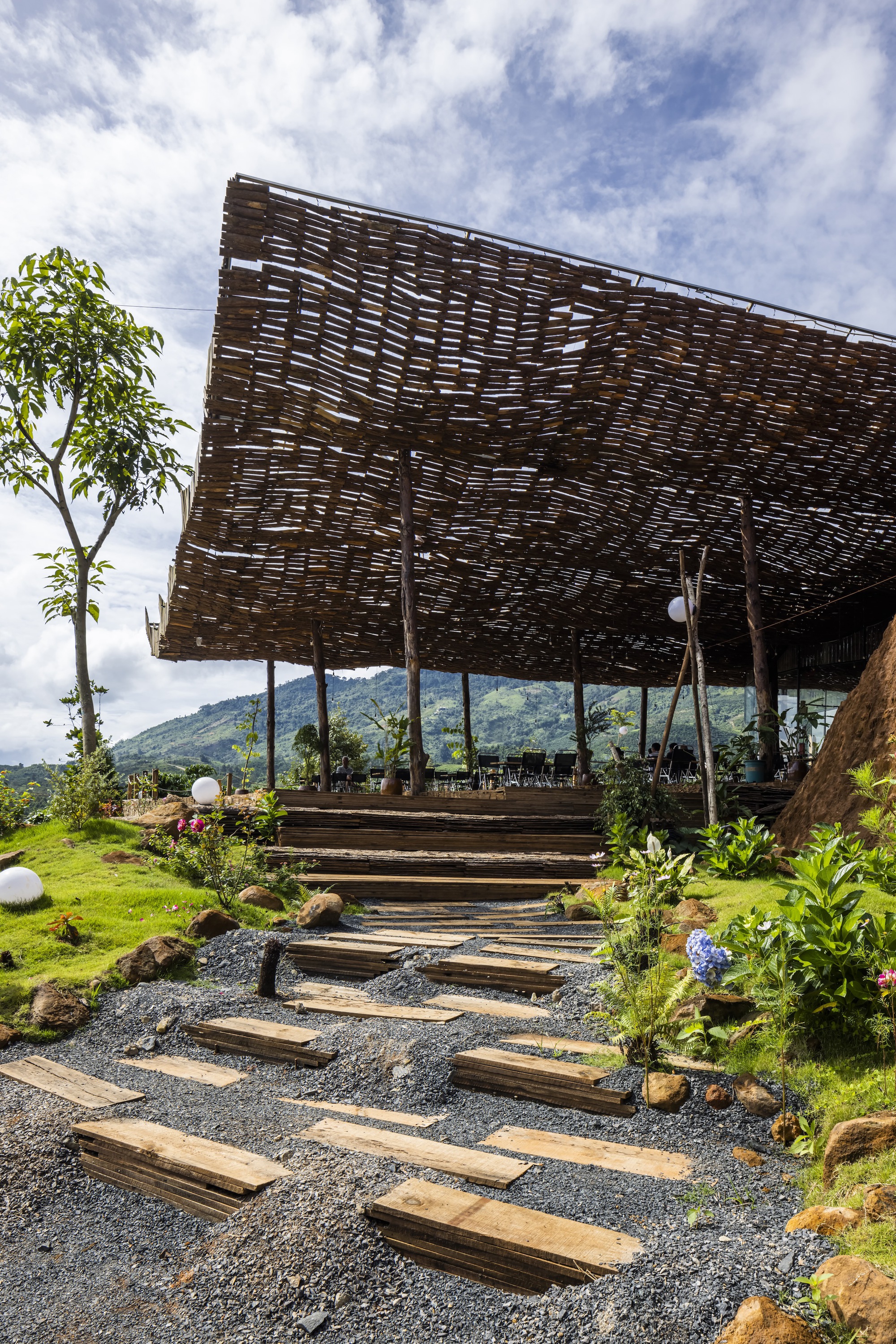 Timber plank pathway on gravel leading toward the woven screen pavilion with hillside landscape beyond