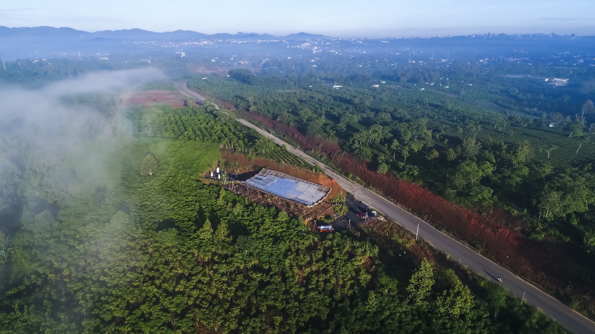 Aerial view of a low-slung roof nestled among agricultural plots and forested hillsides in morning mist