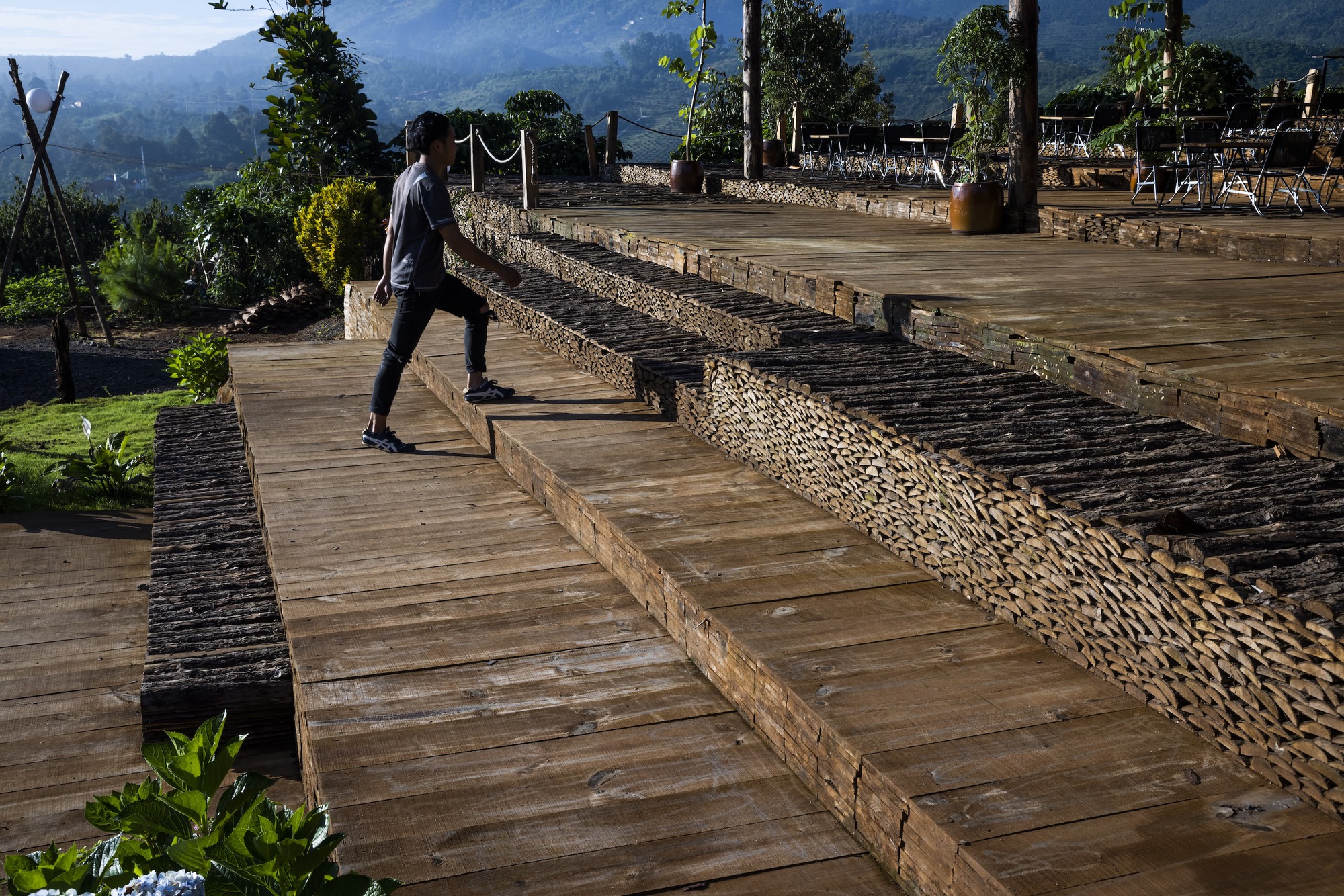 Visitor crossing wide timber deck platforms bordered by woven bamboo retaining walls in morning light