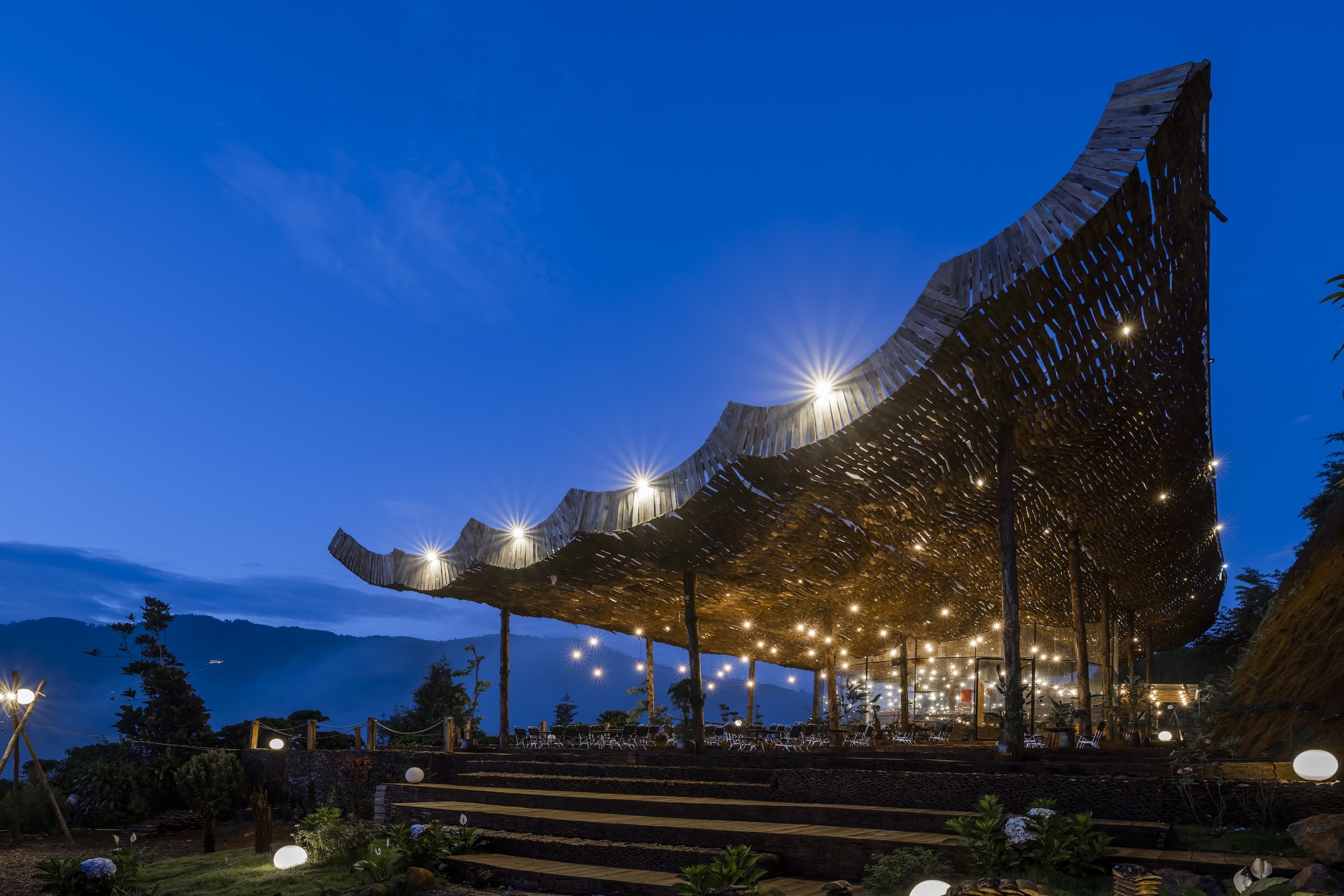 Woven timber canopy with undulating edge and integrated lighting above terraced timber seating at twilight