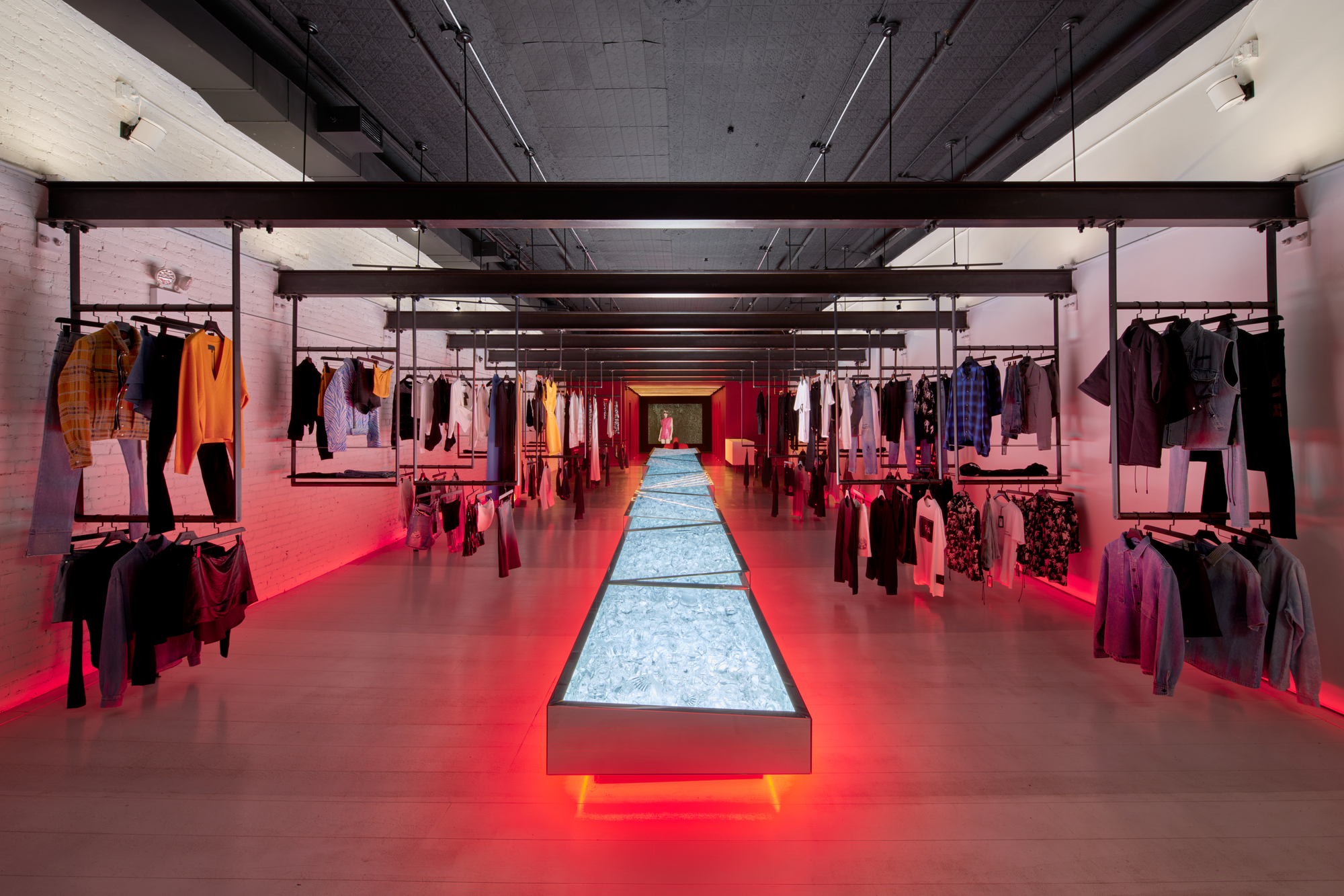 Long interior view down a central illuminated display table flanked by suspended clothing racks and exposed ceiling beams