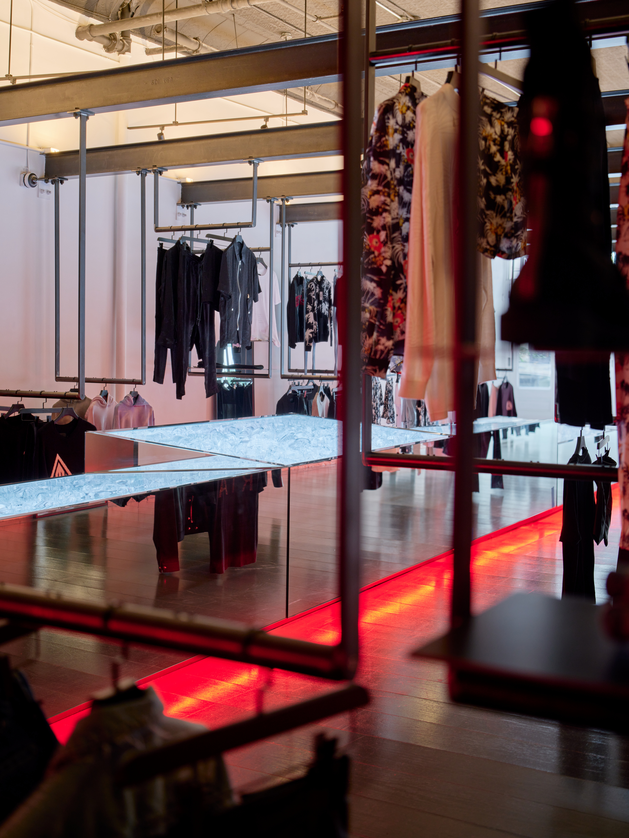 Retail interior with steel clothing racks, marble display tables, and red floor lighting beneath dark wood flooring