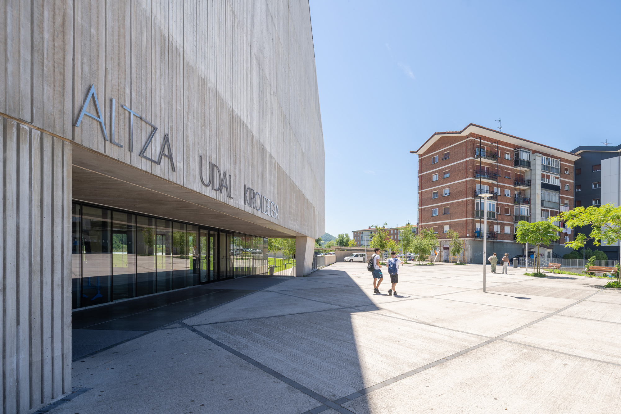 Vertically ribbed concrete facade with recessed entrance and pedestrians crossing the plaza under blue sky