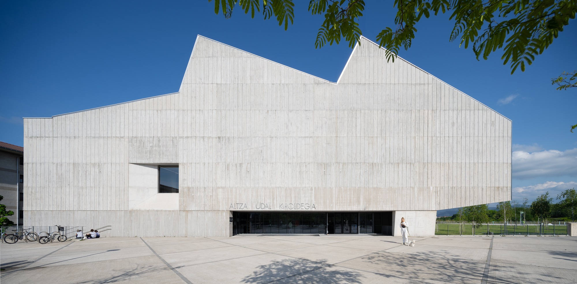 Ribbed concrete facade with angular roofline and horizontal entrance slot beneath tree branches