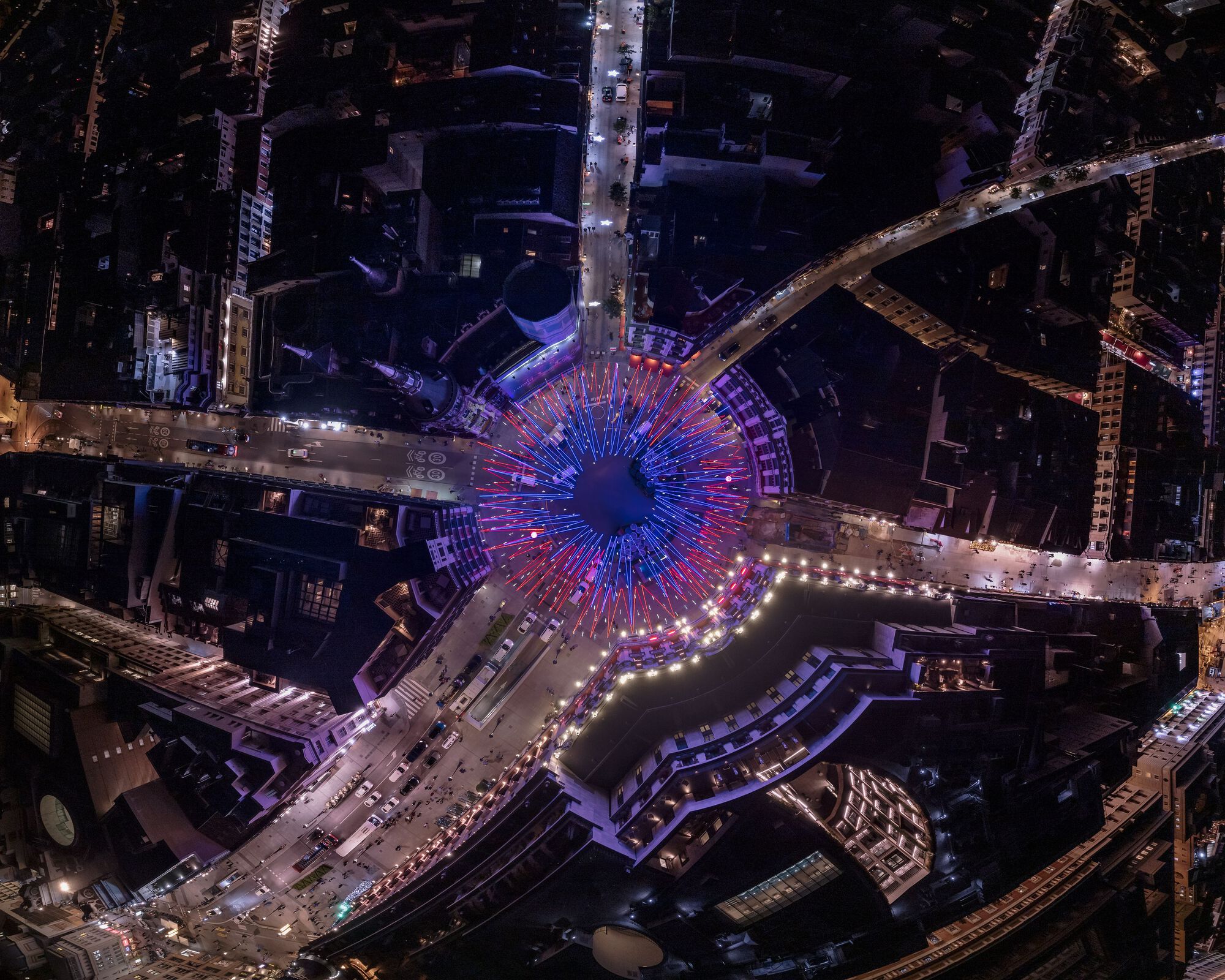 Aerial view of circular light installation with radiating blue and red tubes extending into surrounding streets