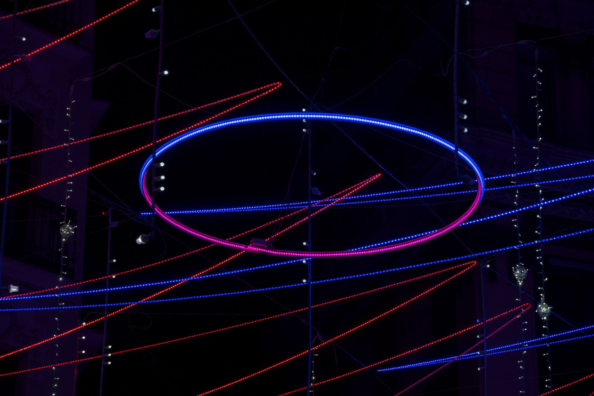 Illuminated elliptical ring with blue and pink LED lights suspended among horizontal light strands at night