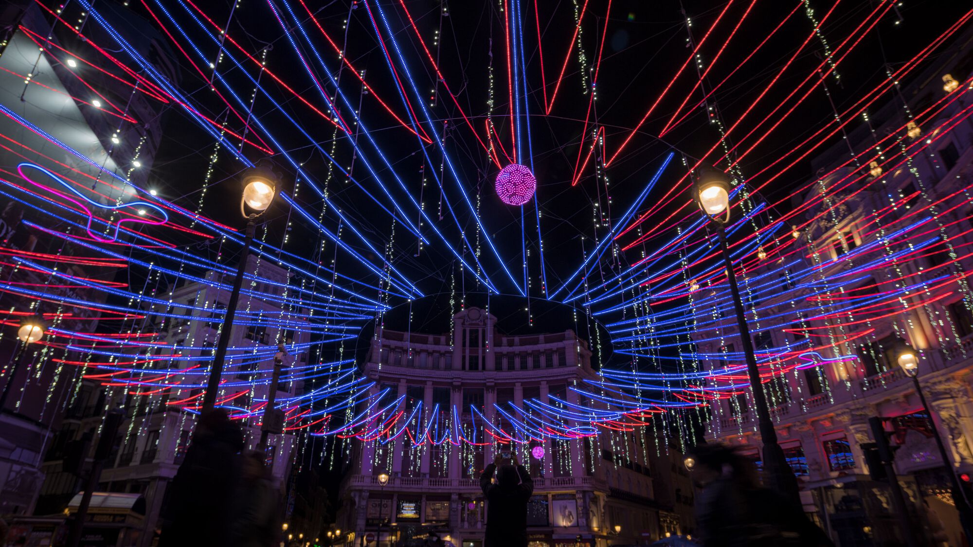 Radial array of suspended LED light tubes converging at a central pendant sphere above surrounding historic facades