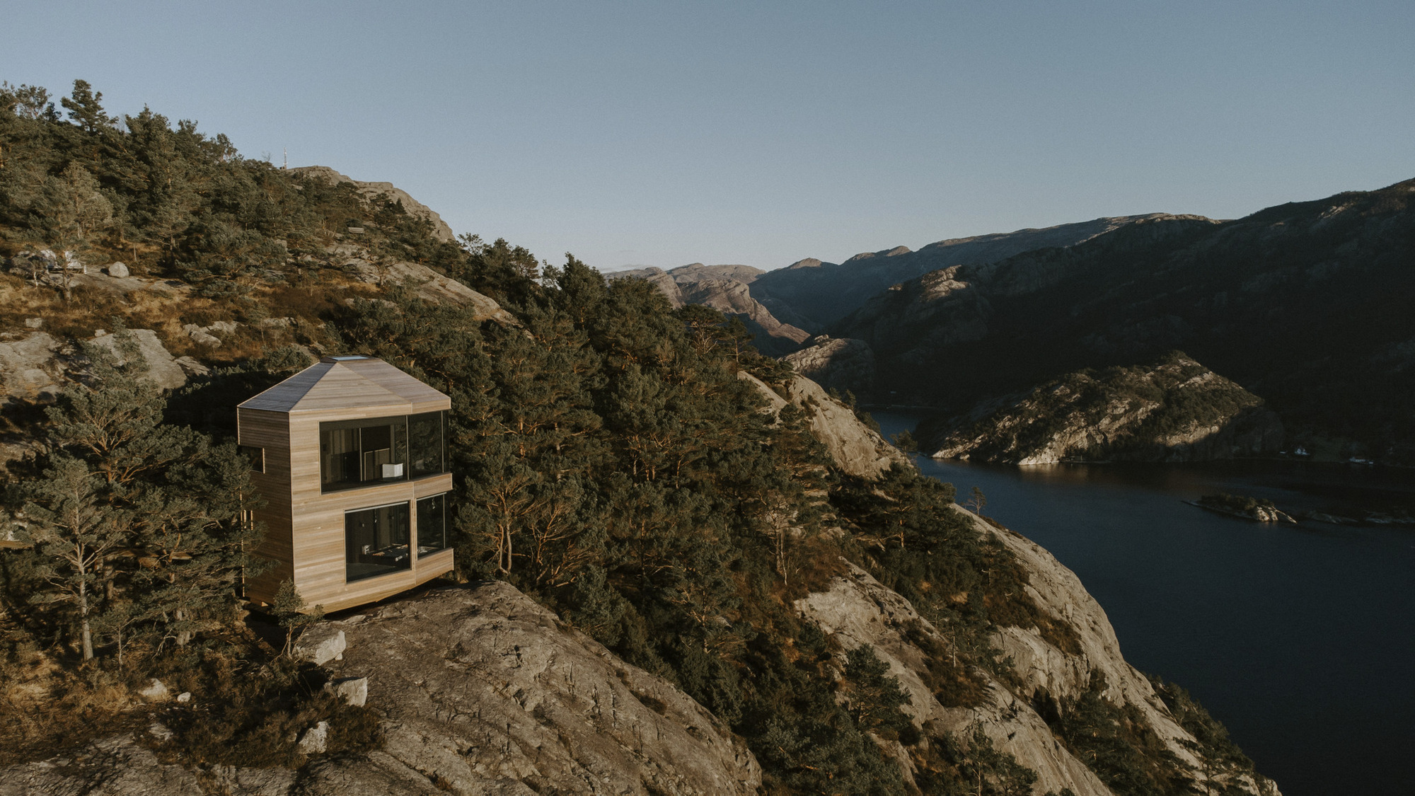 Two-story timber-clad cabin perched on a rocky outcrop above a fjord in late afternoon light