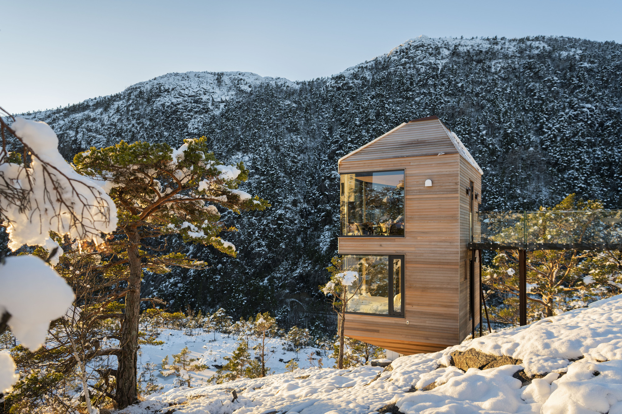 Hexagonal timber-clad structure with floor-to-ceiling glazing set among snowy pines and rock outcrops