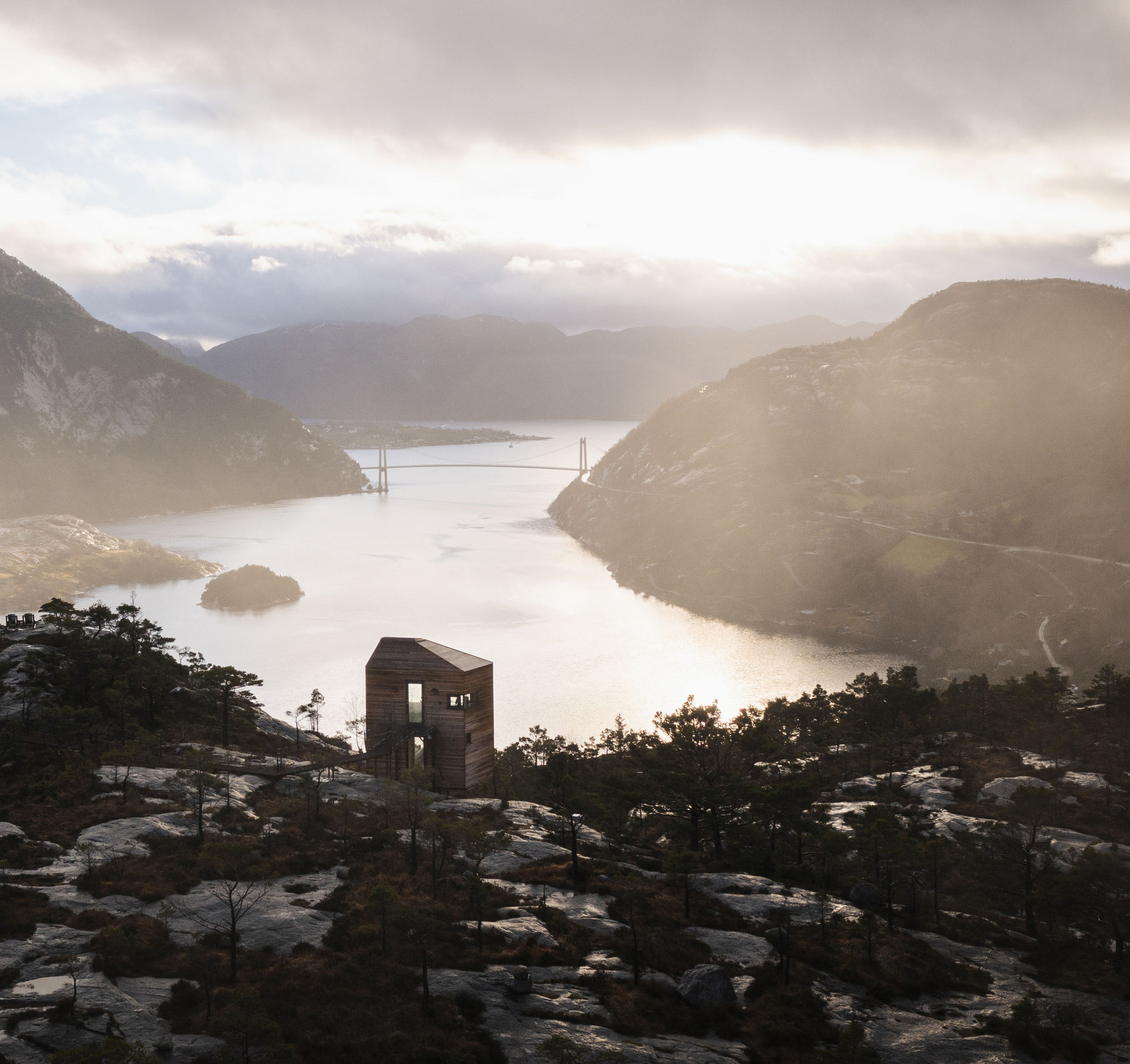 Timber cabin on a hillside overlooking a misty fjord with a suspension bridge at sunrise
