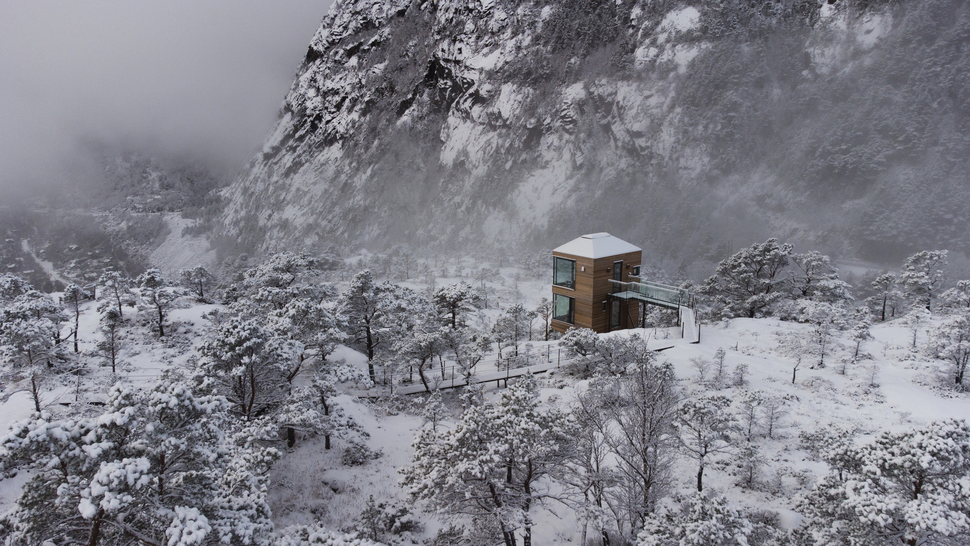 Drone view of the timber cabin in a snowy clearing against a rocky cliff in fog