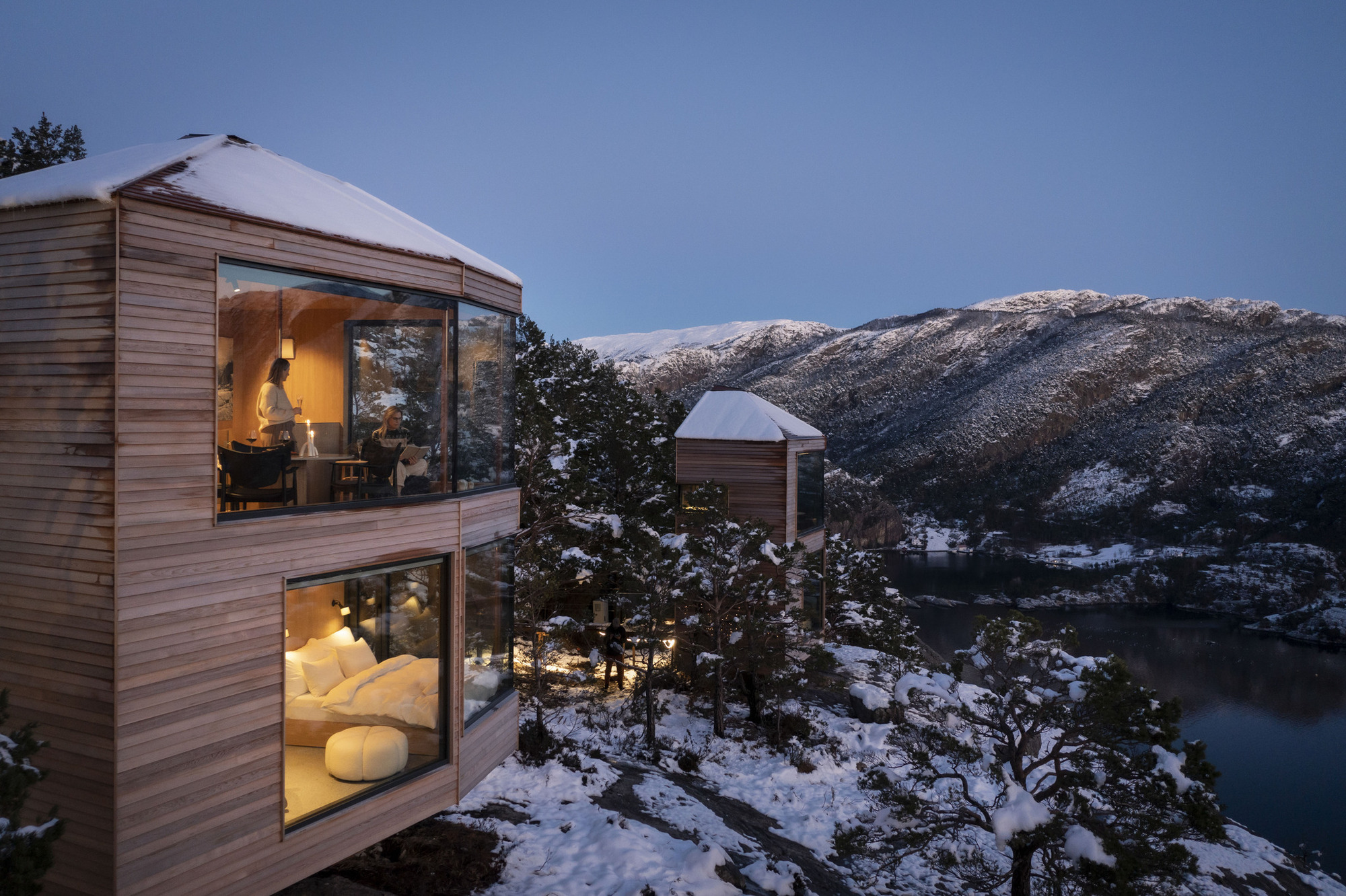 Two-storey facade with full-height glazing and timber cladding overlooking a snowy fjord at dusk