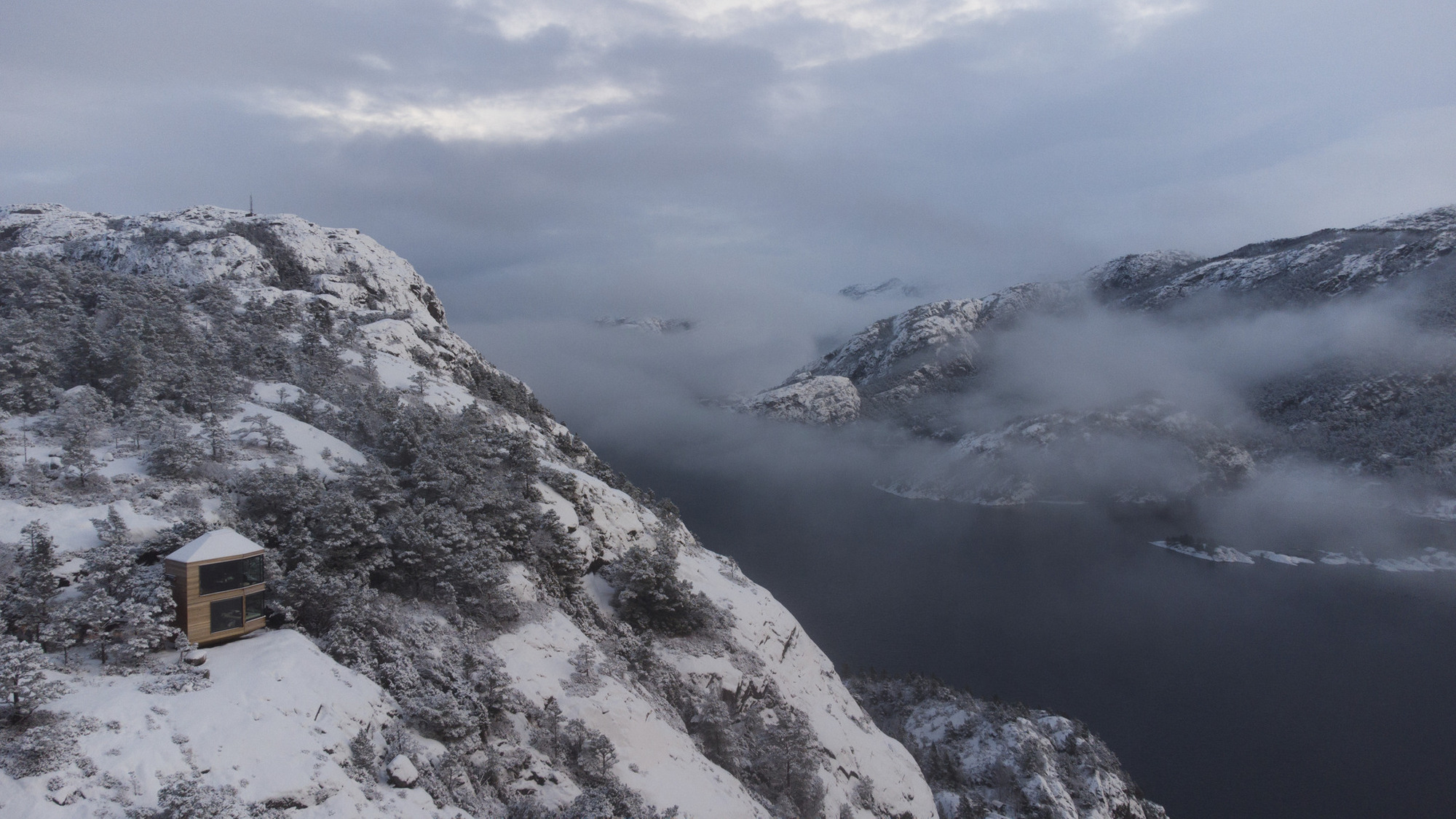 Distant view of the cabin on a snow-dusted slope above a misty fjord