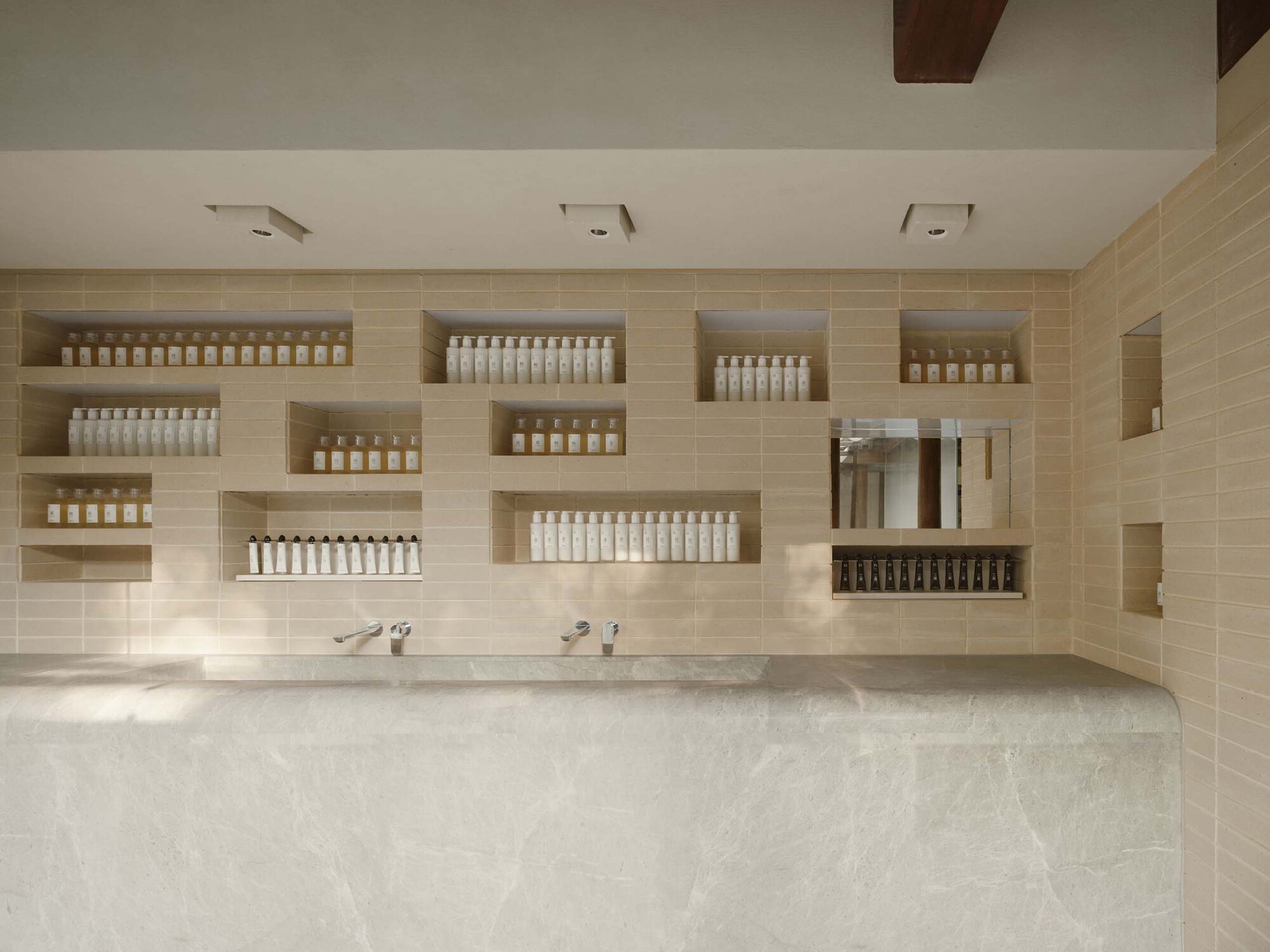 This architectural image showcases a modern bathroom interior with a minimalist design. The space features a beige tile wall with recessed shelves displaying va