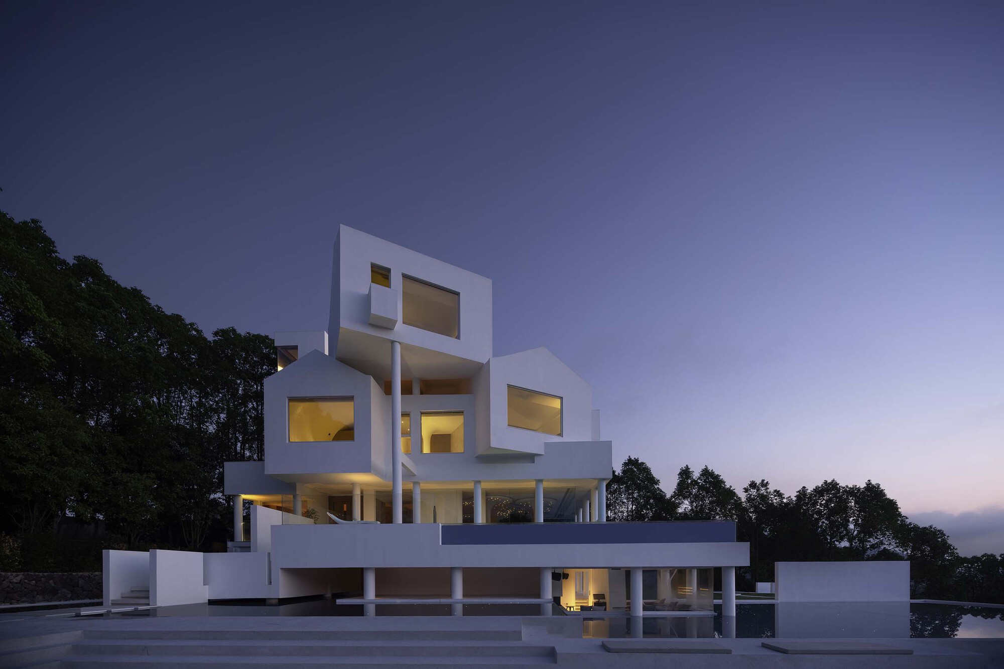 White stacked volumes with illuminated rectangular window openings at dusk beside a reflecting pool