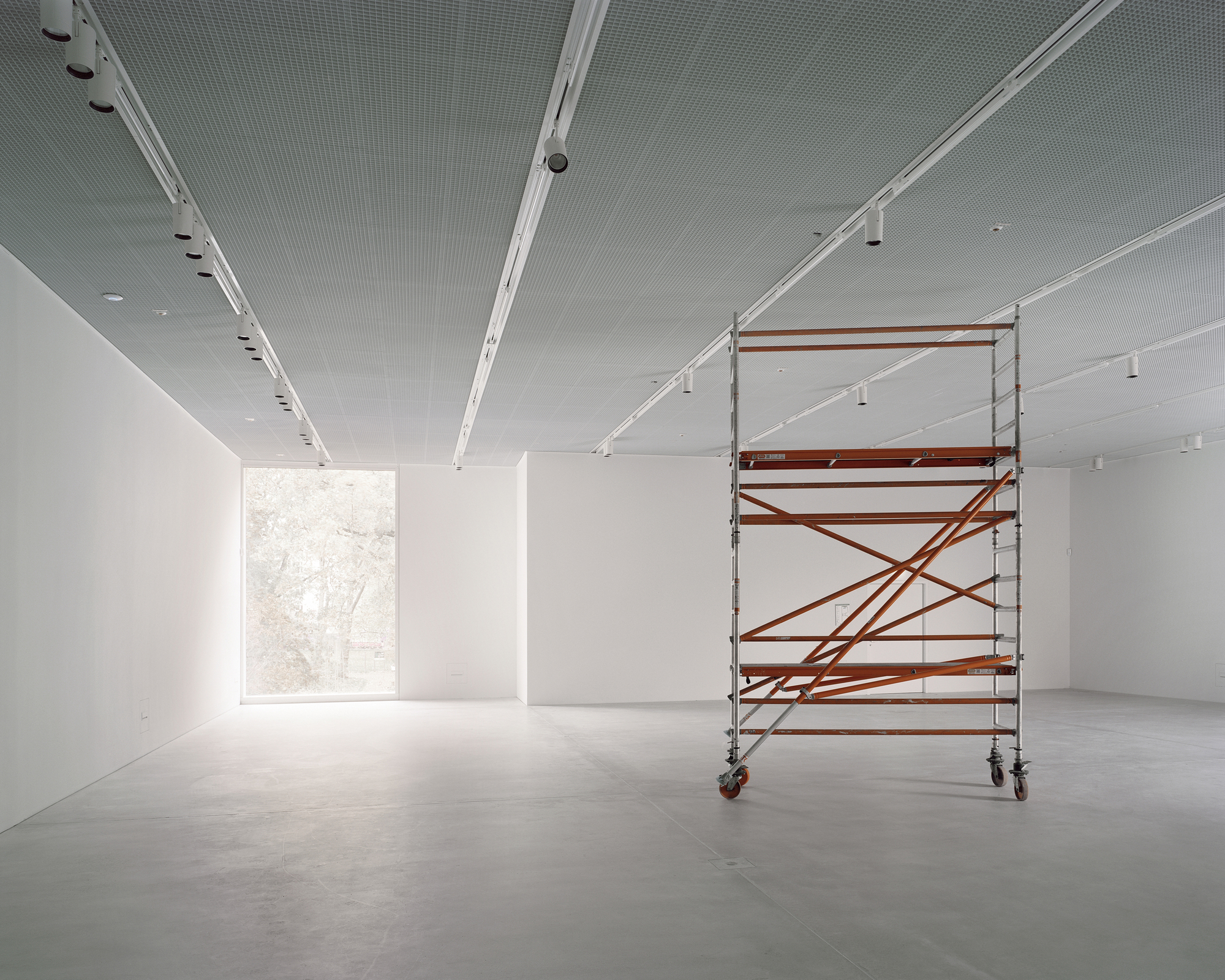 White gallery space with polished concrete floor, track lighting, and a rolling scaffold near the window