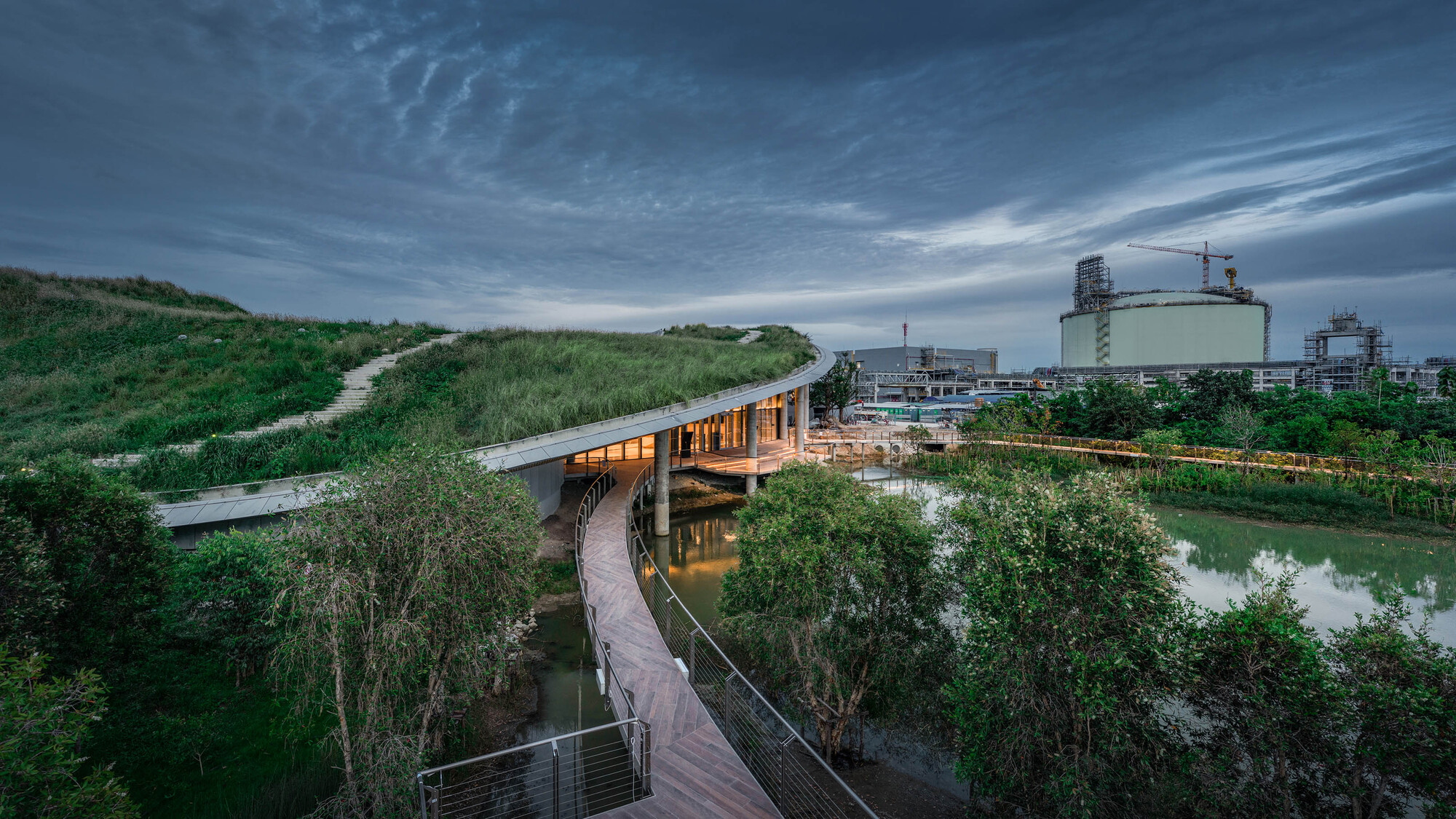 Elevated walkway bridging a planted wetland and pond at dusk with industrial tanks in the distance