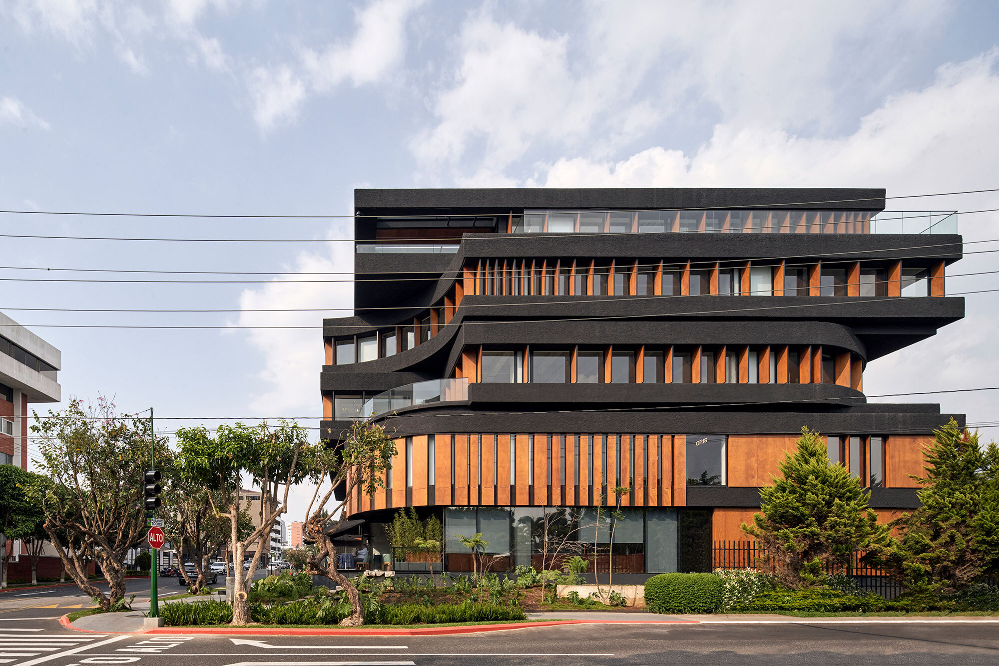 Stacked facade with dark concrete slabs and vertical timber screening viewed from the street under cloudy skies