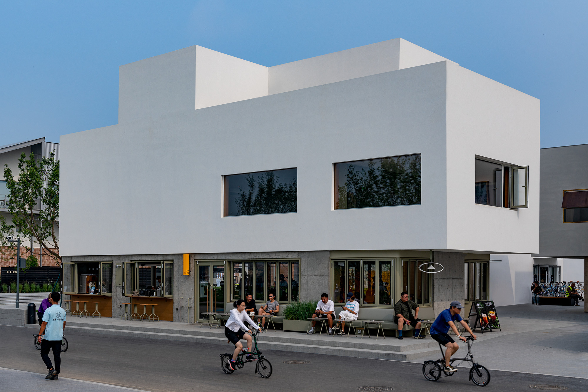 Street view of the white stucco facade with ground-floor cafe and pedestrians on bicycles at dusk