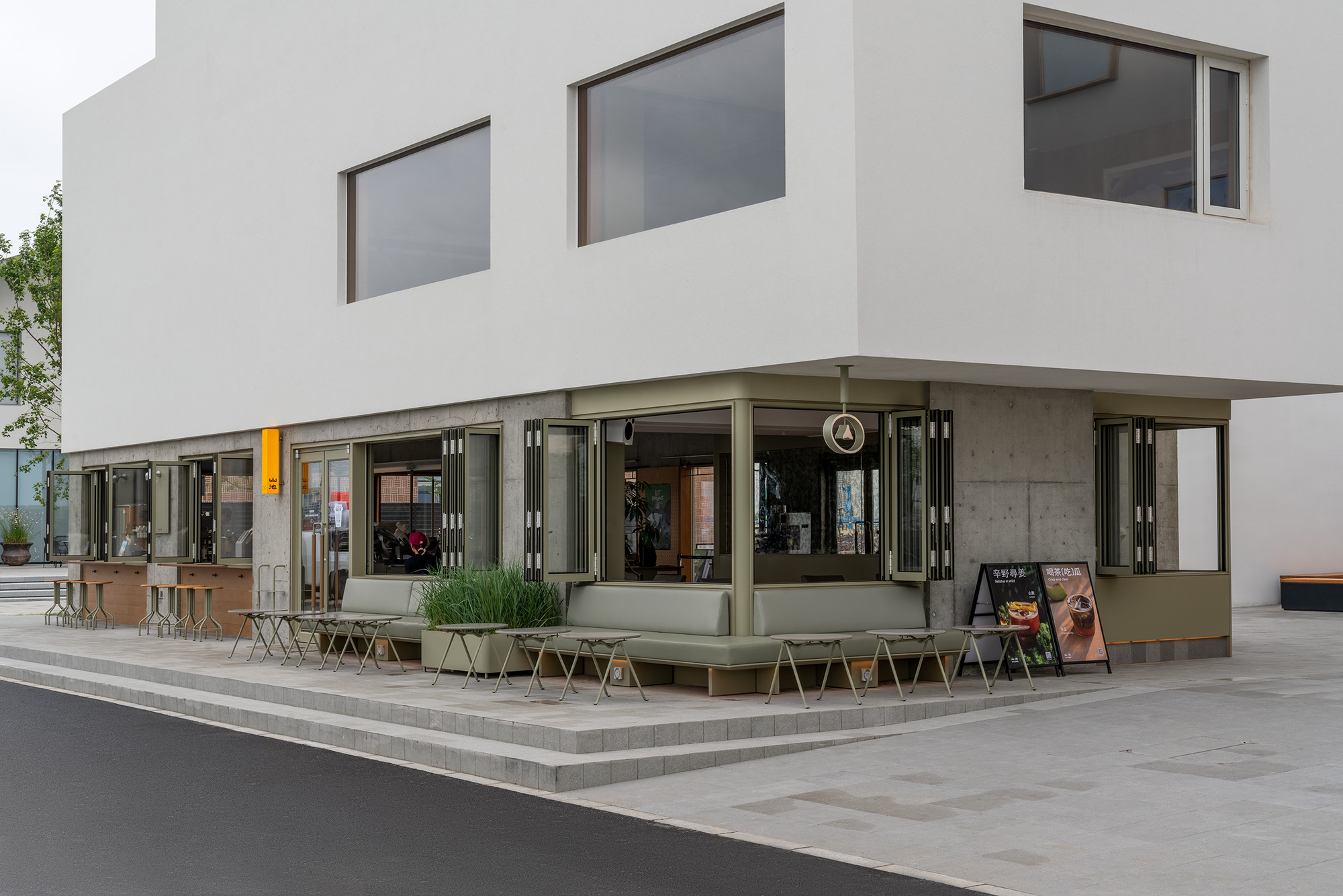 Corner detail of the folding glass storefront with olive-green frames and outdoor seating on the plaza