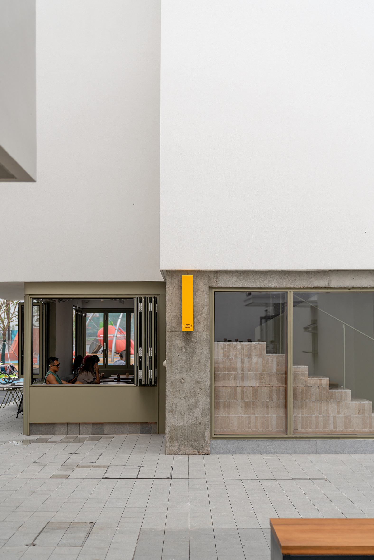 Ground-level exterior showing the folding cafe window, yellow column, and interior staircase beyond glazed entry