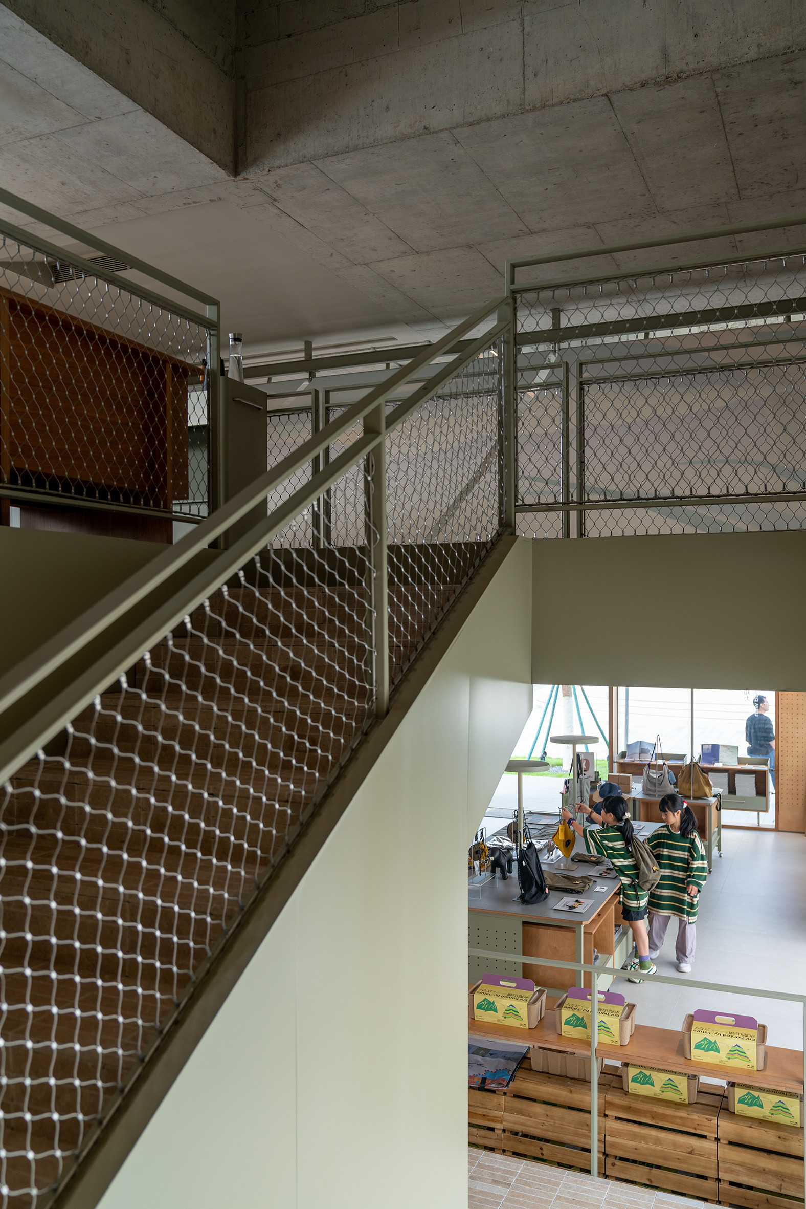 Staircase with mesh balustrades and exposed concrete ceiling overlooking a children's play area below