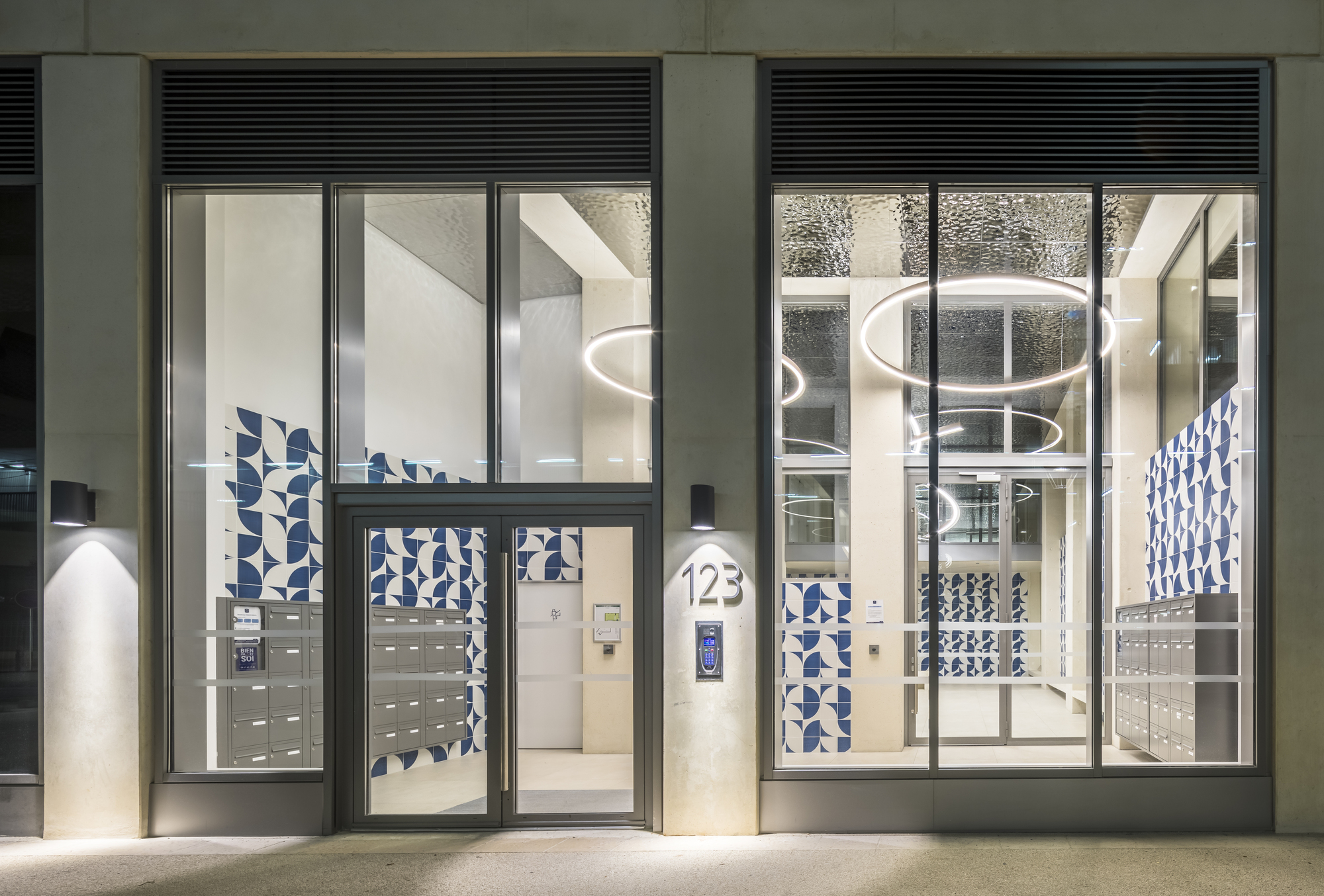 Glass entry lobby doors with integrated letterboxes and blue patterned tile panels at night