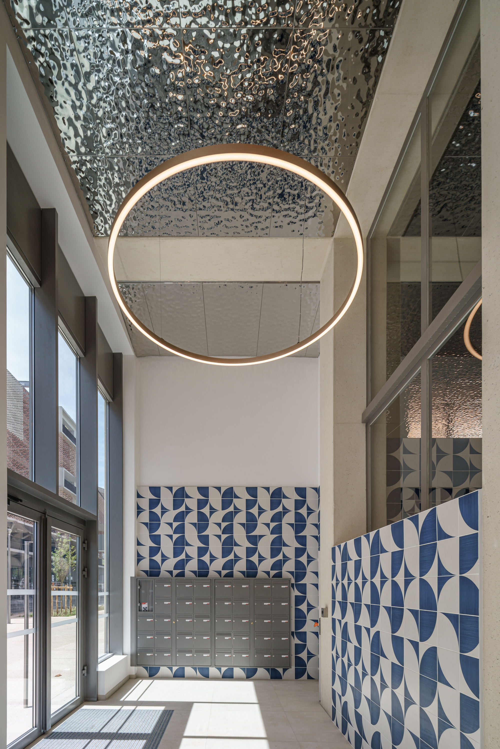 Double-height lobby with circular brass pendant light beneath a perforated metal ceiling and blue geometric tiled wall