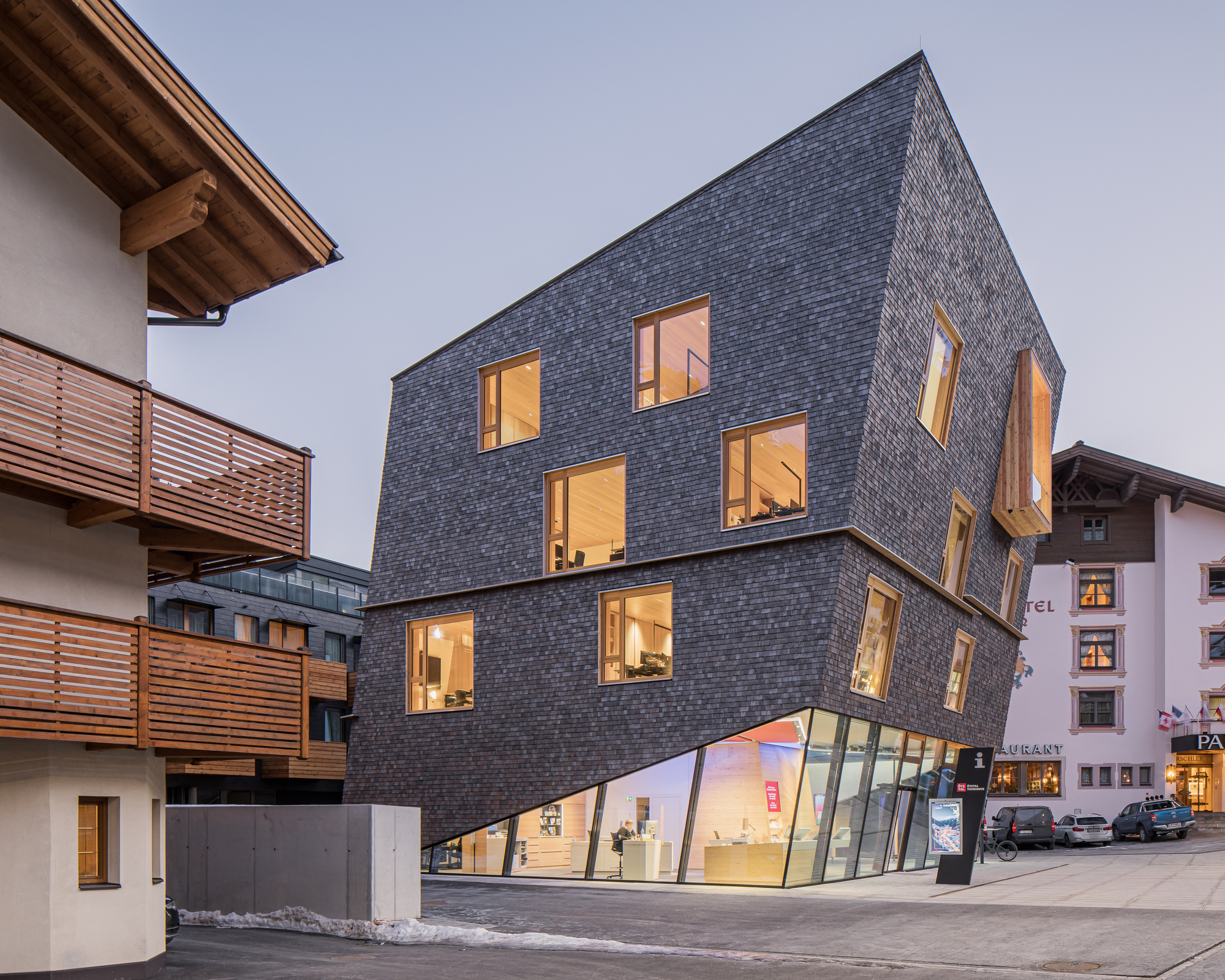 Three-story shingled volume with illuminated windows and an angled glass storefront at dusk