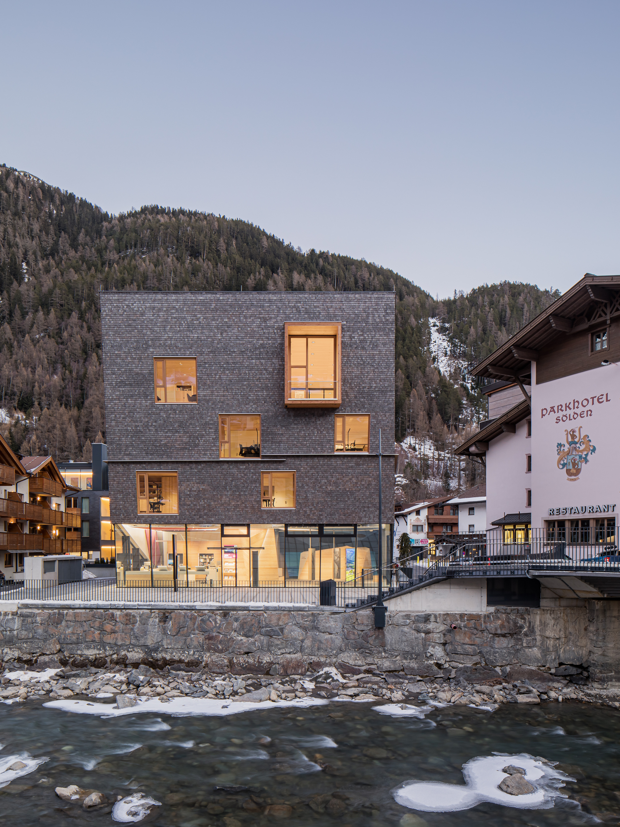 Dark slate shingle facade with illuminated windows overlooking a rushing river at dusk